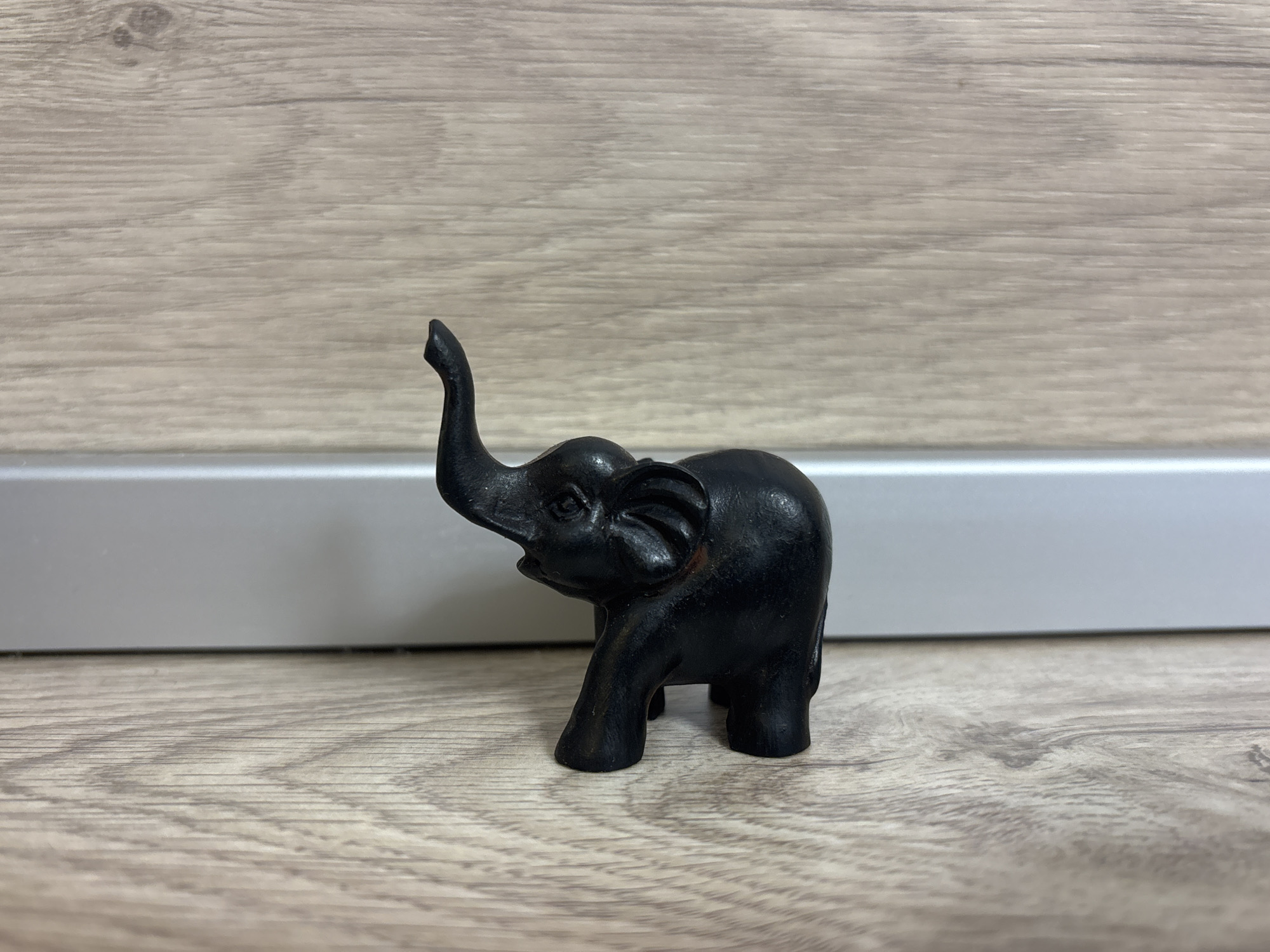 A black elephant figurine stands on a light wood-grain surface in front of a gray and white wall. Unknown, Unknown is not part of the details in the image.
