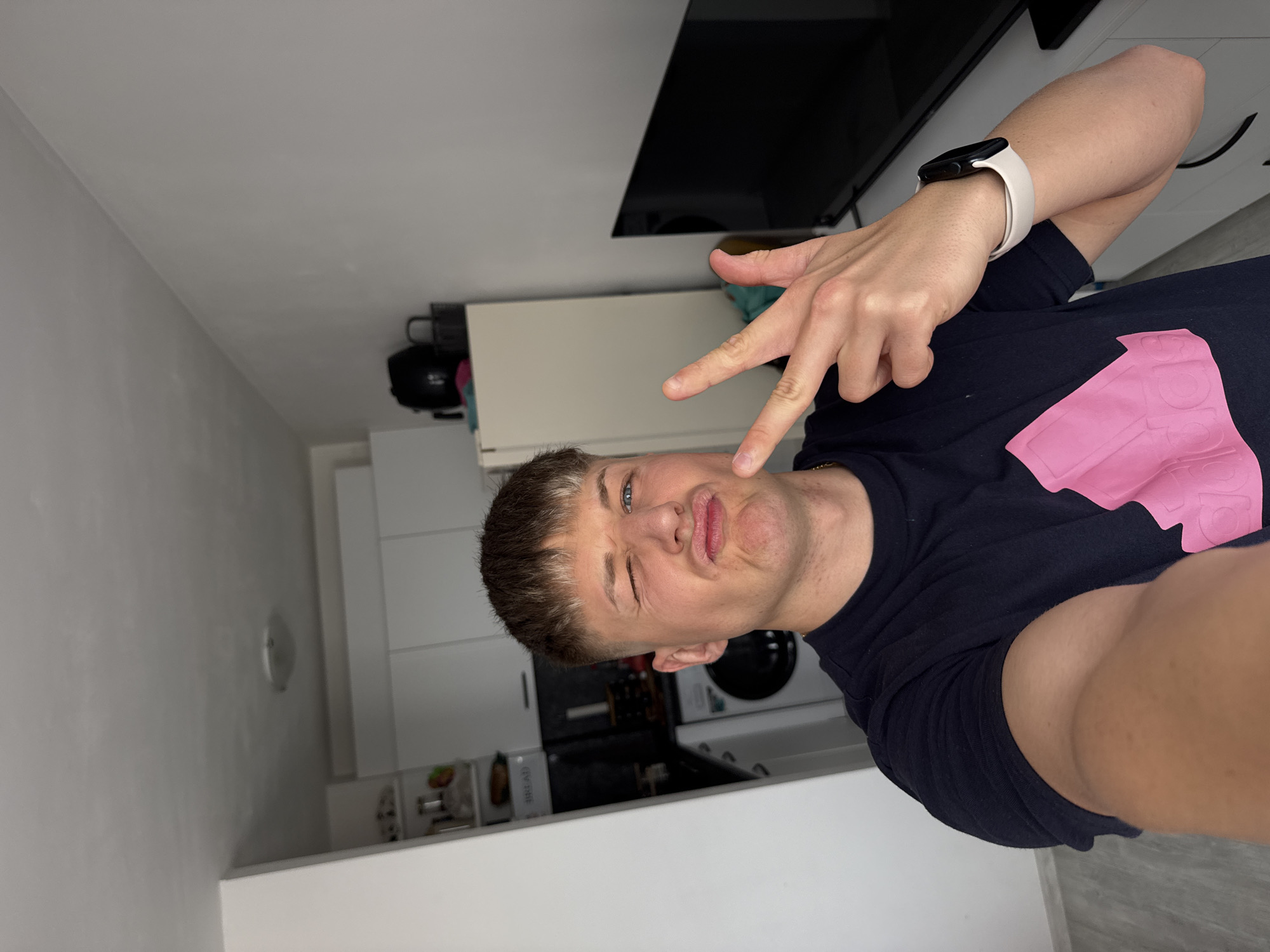 A young man is taking a selfie indoors. He is wearing a dark blue t-shirt with a pink design and a white smartwatch. He is making a peace sign with his right hand and a funny face. Behind him is a kitchen with white cabinets, a black microwave, and a television mounted on the wall. Based on the style and architecture of the interior, and lacking more specific details, it is difficult to pinpoint the exact location in Plovdiv, Bulgaria.