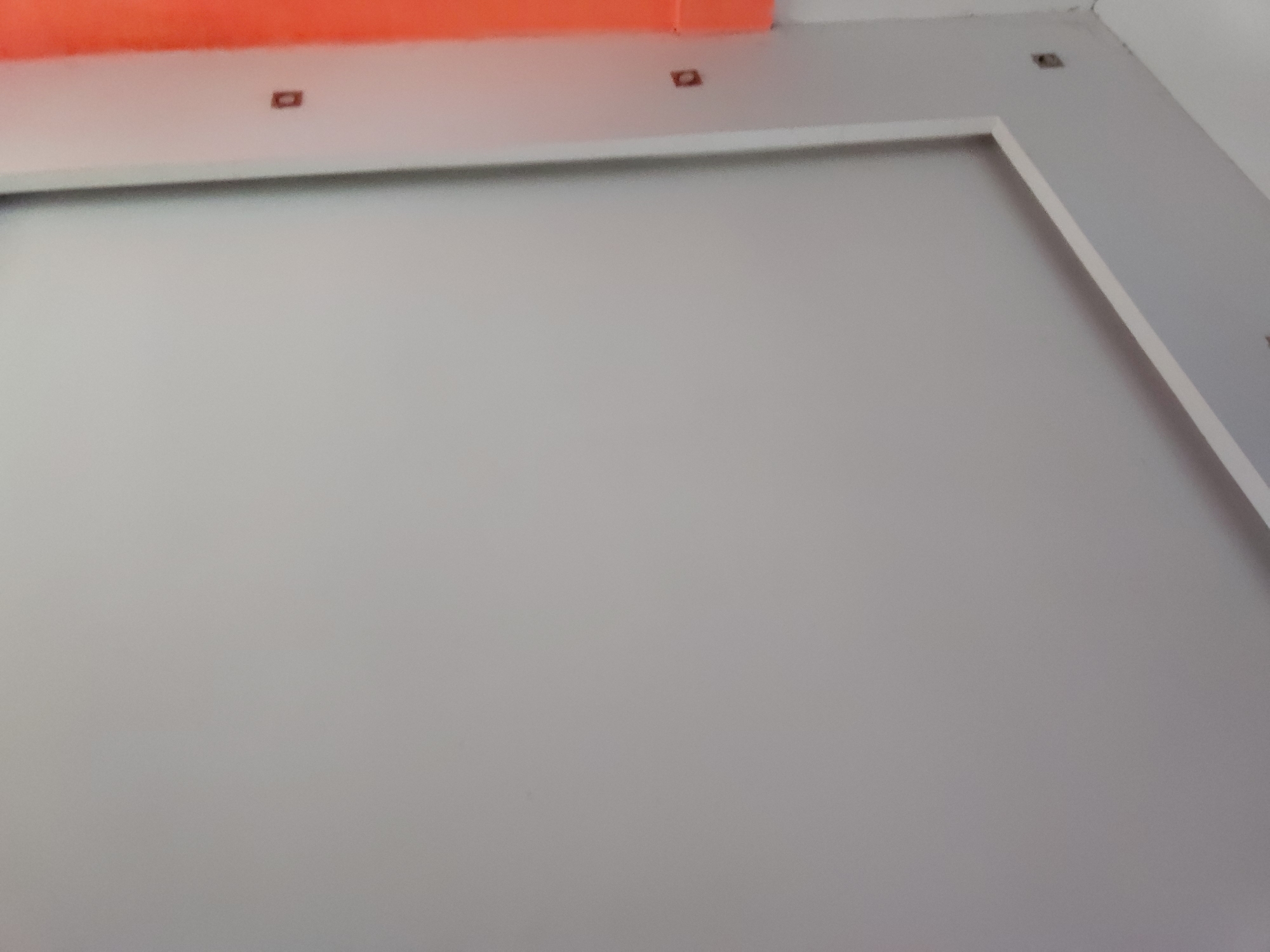 The image captures an indoor architectural detail, primarily focusing on a light gray wall with distinct white and orange elements. The perspective is angled upwards, showing a ceiling or upper wall section.
A broad, horizontal strip of bright orange material, possibly a ceiling beam, decorative panel, or a portion of another wall, is visible at the very top of the frame. Directly below this orange section is a flat, off-white surface. Embedded within this white surface are small, dark reddish-brown, rectangular elements, likely recessed light fixtures or electrical outlets; two are clearly visible on the left and one is partially seen on the right.
The main wall area is a uniform light gray. A prominent feature is a wide, white rectangular frame or trim applied to this gray wall, creating an architectural panel effect. The frame extends along the upper and right edges of the visible wall section.
The lighting appears even, suggesting an interior space with ambient or indirect light. There are no people, visible activities, or discernible text in the image. While the location is provided as Santa Ana, Venezuela, the visual elements themselves are generic and do not offer specific geographical cues. The scene suggests a modern, clean, and perhaps unfinished or newly constructed interior environment.