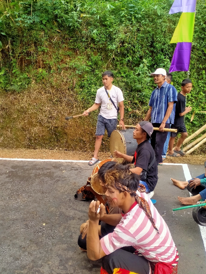 The image captures an outdoor scene in Ngargoyoso, Indonesia, featuring six individuals on and beside a paved surface with a distinct white line. The background consists of a dense, green hillside with varied foliage and some dried vegetation.
In the immediate foreground, two men are seated directly on the pavement. The man closer to the viewer, positioned on the right, wears a red and white striped shirt, black pants, and a patterned headband. He has white paint or markings on his right cheek and holds a cigarette between the fingers of his raised right hand. To his left, another seated man, largely obscured, appears to be holding a small, brown drum (kendang). Partial views of bare feet belonging to an additional seated person are visible further to the right.
In the midground, a man wearing a black t-shirt and a grey baseball cap is seated on a low white object, holding a large drum (gendang or bedug) between his legs, with a drumstick in his right hand. To his right, a standing man in a white t-shirt, blue shorts, and flip-flops holds a large drumstick in his right hand, extending it forward. A black lanyard with an ID card hangs around his neck. Behind him, another standing man wears a blue and white striped shirt and a white baseball cap, holding a long bamboo pole horizontally. To the far right, a young boy in a black t-shirt stands. Several vertical bamboo poles are partially visible on the right side of the frame. Above the standing figures, colorful flags, including purple and lime green, are strung up against the hillside foliage.