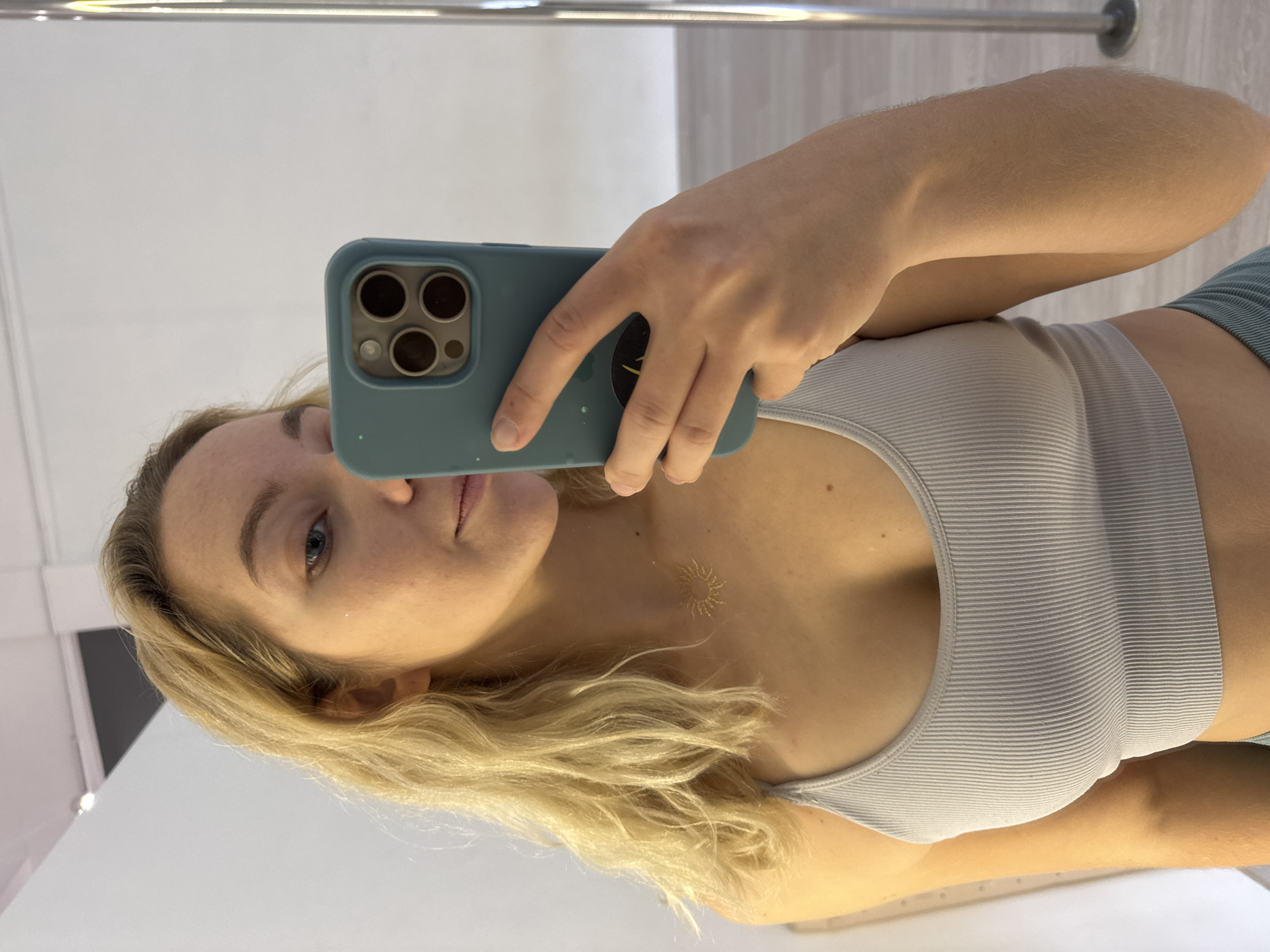 A woman with blonde hair, wearing a grey ribbed tank top and a sun necklace, takes a mirror selfie indoors. She is holding a teal phone in her right hand, which is partially visible. The background appears to be a brightly lit room with white walls and a reflective surface, possibly a mirror. A metal pole is visible on the right side of the frame. There is no indication of the city of Minsk, Belarus.