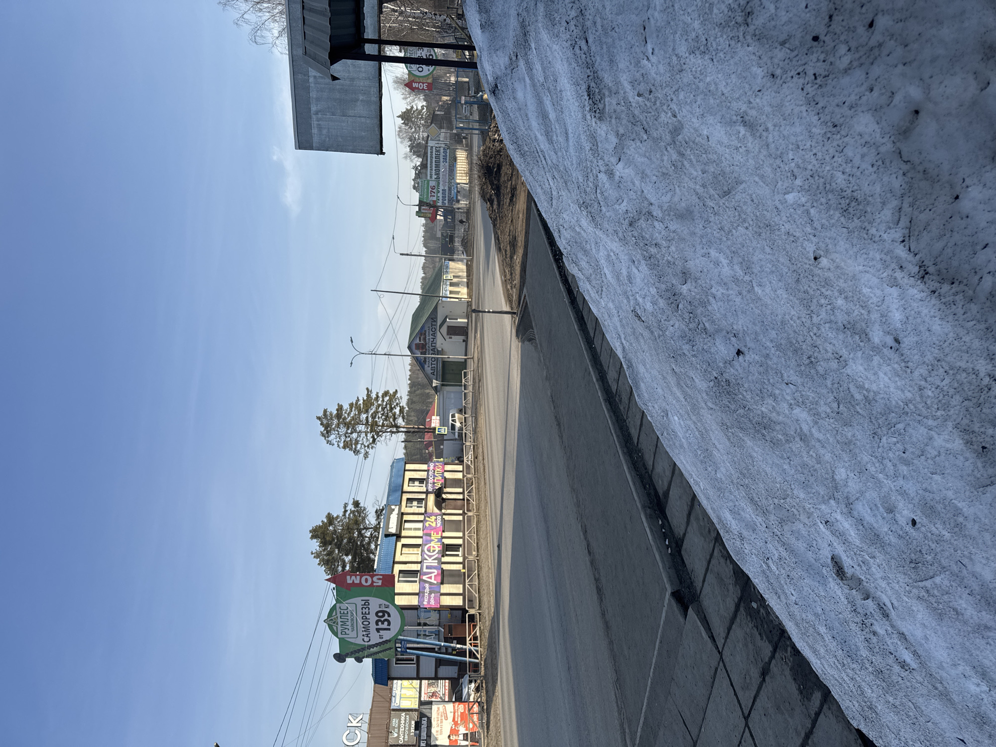 The image shows a street in Chaikovsky, Russia. Along the street are various advertisements, including one for "РУМЛЕС ЧАЙКОВСКИЙ" (Rumles Chaikovsky) offering "САМОРЕЗЫ" (screws) from 139 rubles. Other advertisements include "АЛКОМЕ 24" (Alcome 24), indicating round-the-clock alcohol sales, and "АВТОЗАПЧАСТИ" (auto parts). A snowdrift is visible along the right edge of the street. Buildings are present along the street, and utility poles line the road. The sky is clear and blue.
