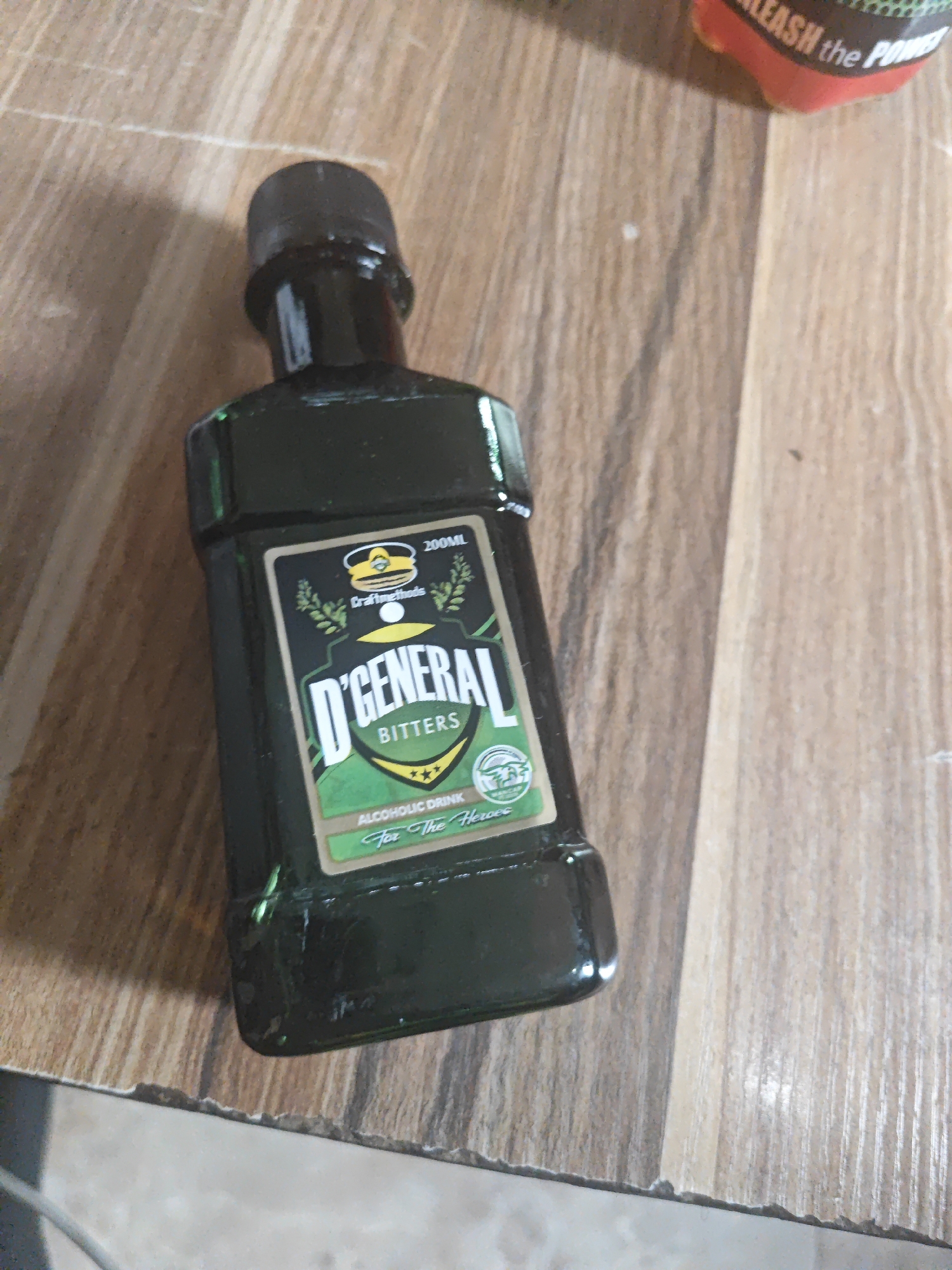 The image shows a dark green glass bottle of D'General Bitters, an alcoholic drink, placed on a wooden surface. The bottle has a black cap and a label with a green and white design. The label features a crest with a soldier's hat and the text "Crafting Methods". Below this, "D'GENERAL" is written in large white letters, followed by "BITTERS" in smaller, black text. There are also decorative sprigs of leaves and a small shield with three stars. At the bottom of the label, "ALCOHOLIC DRINK" and "For The Heroes" are visible. The bottle is the main subject, positioned slightly to the left of the frame. The background is a wooden surface with a visible grain pattern. A blurry object with red and black text, possibly another bottle or packaging, is visible in the upper right corner of the image. The lighting suggests it is daytime, and the overall scene appears to be indoors. No people are visible in the image. The location is identified as Bukuru, Nigeria.