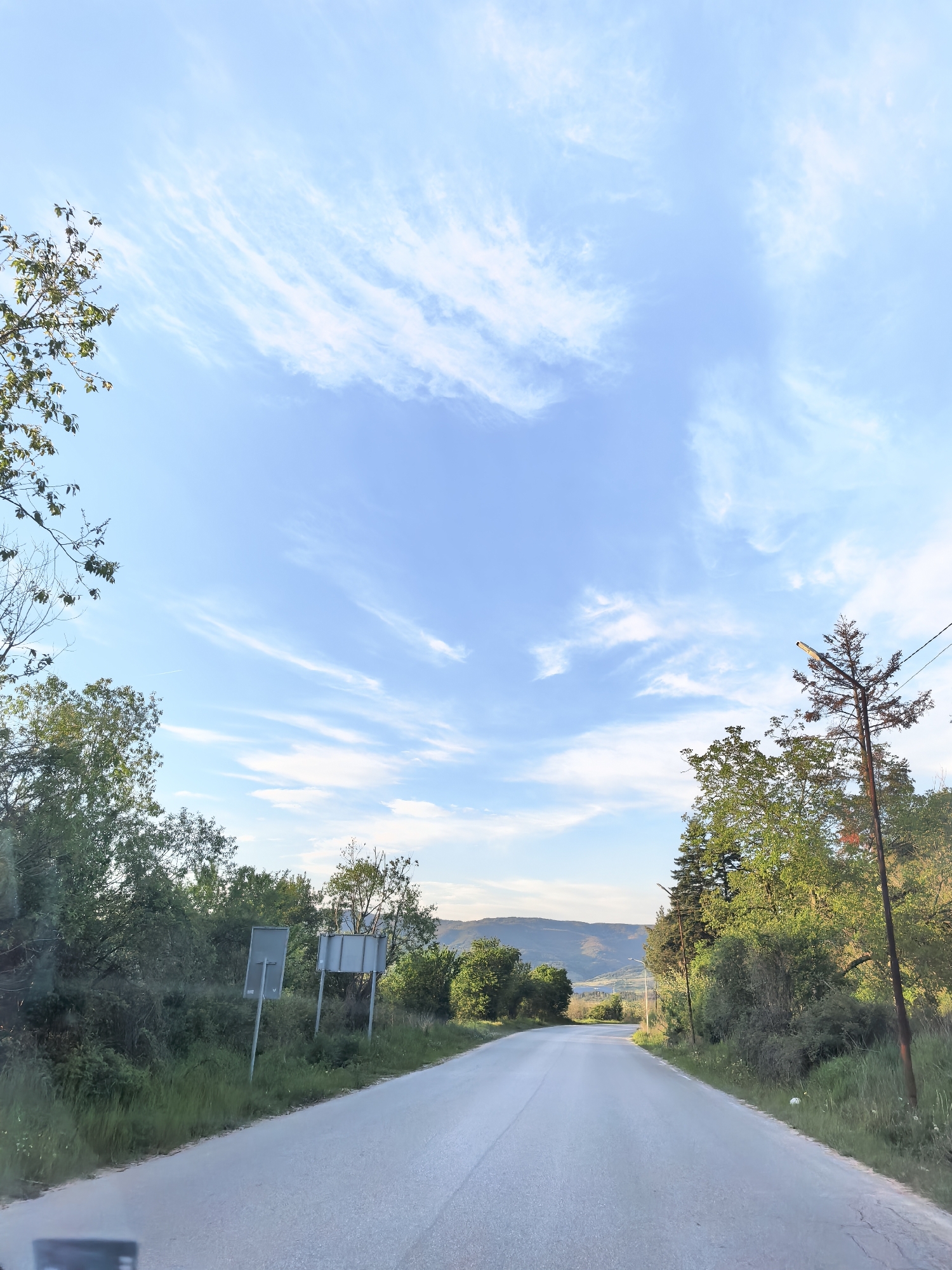 The image depicts a paved road leading into the distance in Sapareva Banya, Bulgaria. The road is bordered by green vegetation and trees on both sides, with mountains visible in the background. The sky is blue with scattered white clouds. Road signs are visible on the left side of the road, partially obscured by vegetation. A utility pole with a light fixture is present on the right side of the road. The lighting suggests it is daytime. The foreground shows a part of the vehicle's dashboard.