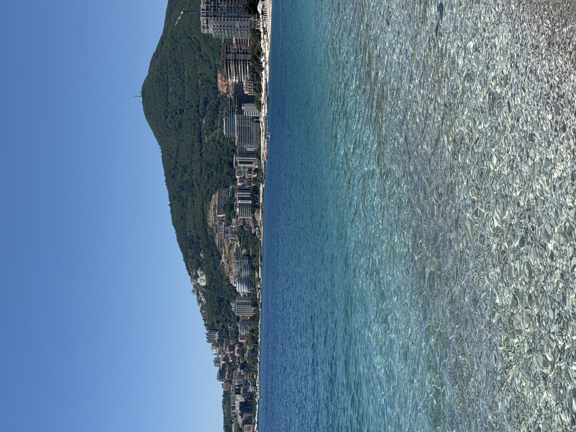 The image shows the shoreline in Budva, Montenegro, with the clear, shallow sea in the foreground transitioning to a deeper blue. The seabed is visible through the clear water. A steep, green hill rises along the shoreline, densely populated with buildings, including hotels and residential structures. Some construction is visible on the hillside. The sky is clear and blue.