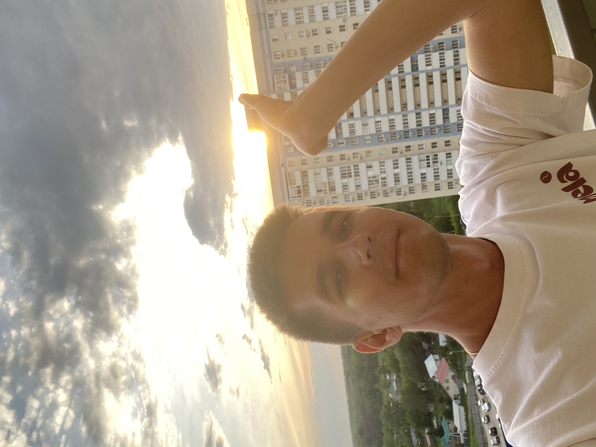 A man stands on a balcony in Novokuybyshevsk, Russia, holding his hand up to block the setting sun. The sky is filled with dramatic clouds, and the sun's rays are visible between them. In the background, a multi-story apartment building dominates the cityscape, with trees and smaller houses visible below. The man is wearing a white t-shirt and a thin gold chain.
