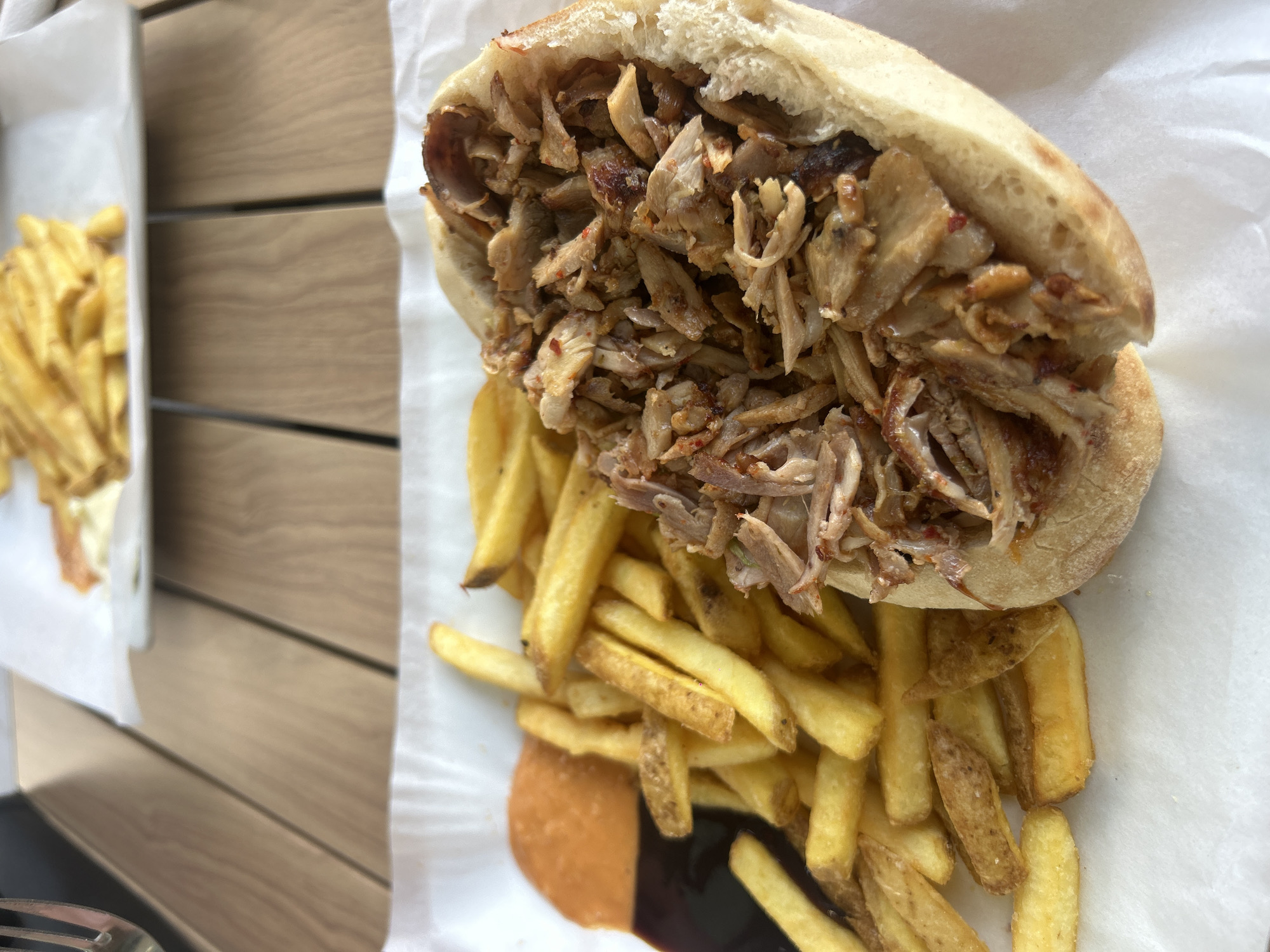 A large, generously portioned meal consisting of a kebab or shawarma sandwich and french fries is depicted in an overhead, slightly angled shot. The sandwich features a split pita bread overflowing with thinly sliced, seasoned cooked meat, likely chicken or lamb, with visible red spice flakes.
A substantial serving of golden-brown french fries is arranged around and partially beneath the sandwich. Two distinct sauces are visible on the white paper lining at the bottom: one light orange/beige and another dark brown/black. The food is presented on white paper, suggesting a casual dining experience, possibly takeaway or fast food.
The setting appears to be a wooden table, with a blurred background showing another plate or container of french fries to the left. The lighting is bright and even, indicating daytime. No people are visible, and there is no discernible text in the image. The scene conveys a hearty, casual meal.