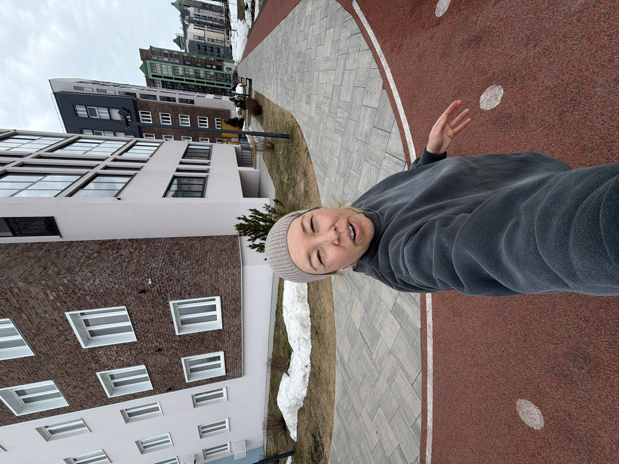 A rotated eye-level outdoor shot depicts a person in a gray beanie and gray hooded sweatshirt standing next to a walkway. The walkway is comprised of gray stone and red rubber. A building is to the left. There is snow on the grass next to the walkway. Other buildings are visible in the distance. The location is the city of Pervomaysky, Russia.