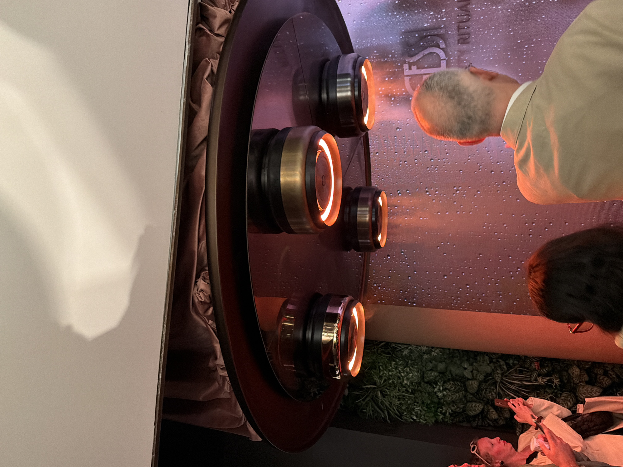 The image depicts an interior scene, possibly an exhibition or a store, where various people are interacting with a display. The display features a circular structure with four cylindrical objects mounted on it, each with a glowing orange light emanating from its center. A person with gray hair wearing a tan jacket is leaning in to observe the display closely, while another individual with dark hair and glasses stands nearby. In the foreground, another person wearing white clothing and sunglasses is seen looking at a phone, possibly interacting with the display or the environment. The backdrop includes a wall with a water droplet pattern and the word "RITUAL" visible, suggesting a theme or concept related to the displayed objects. The environment is further decorated with greenery, contributing to the overall ambiance.