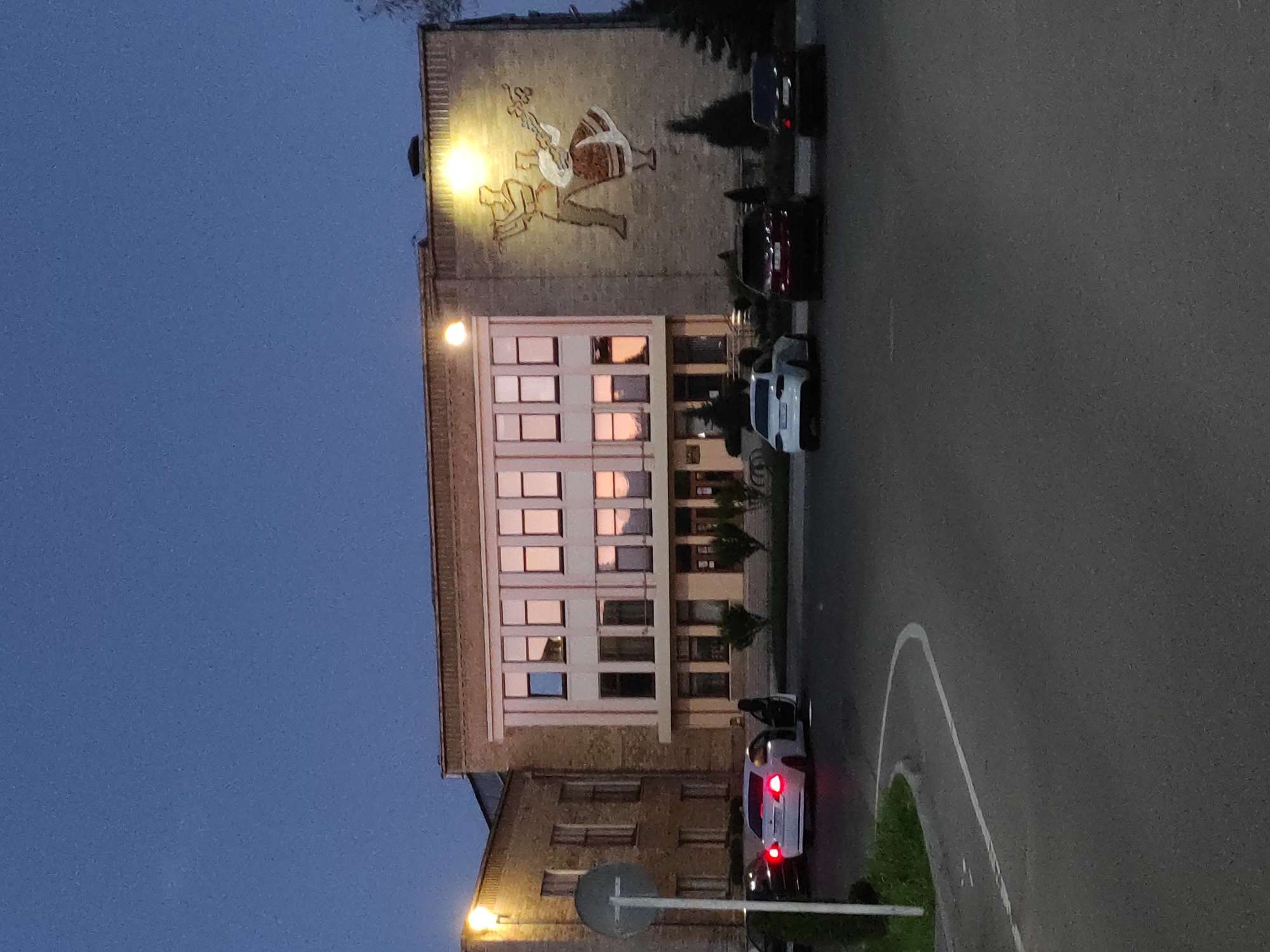 The image captures a building with a mural on its upper section depicting two people, one holding a musical instrument. The building has multiple windows reflecting a sunset or sunrise. Several parked cars are visible on the street beside the building. A street sign is in the foreground. The sky is a dark blue, suggesting dusk or dawn. The location appears to be a city, with the specific city being Unknown, Unknown.