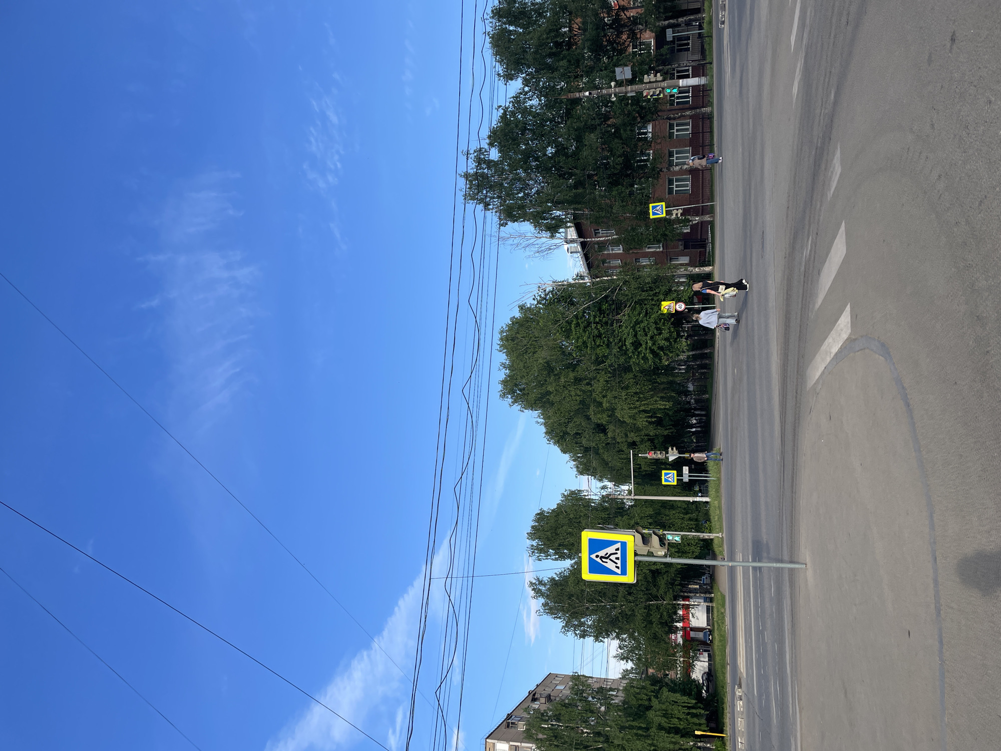 The image shows a street scene in Izhevsk, Russia. The sky is bright blue with wispy clouds, and several electrical wires stretch across the top left of the frame. A street with a crosswalk is visible, with a "pedestrian crossing" sign prominently displayed. Two people are crossing the street with a person lying down on the crosswalk. Buildings with green trees can be seen on both sides of the street.