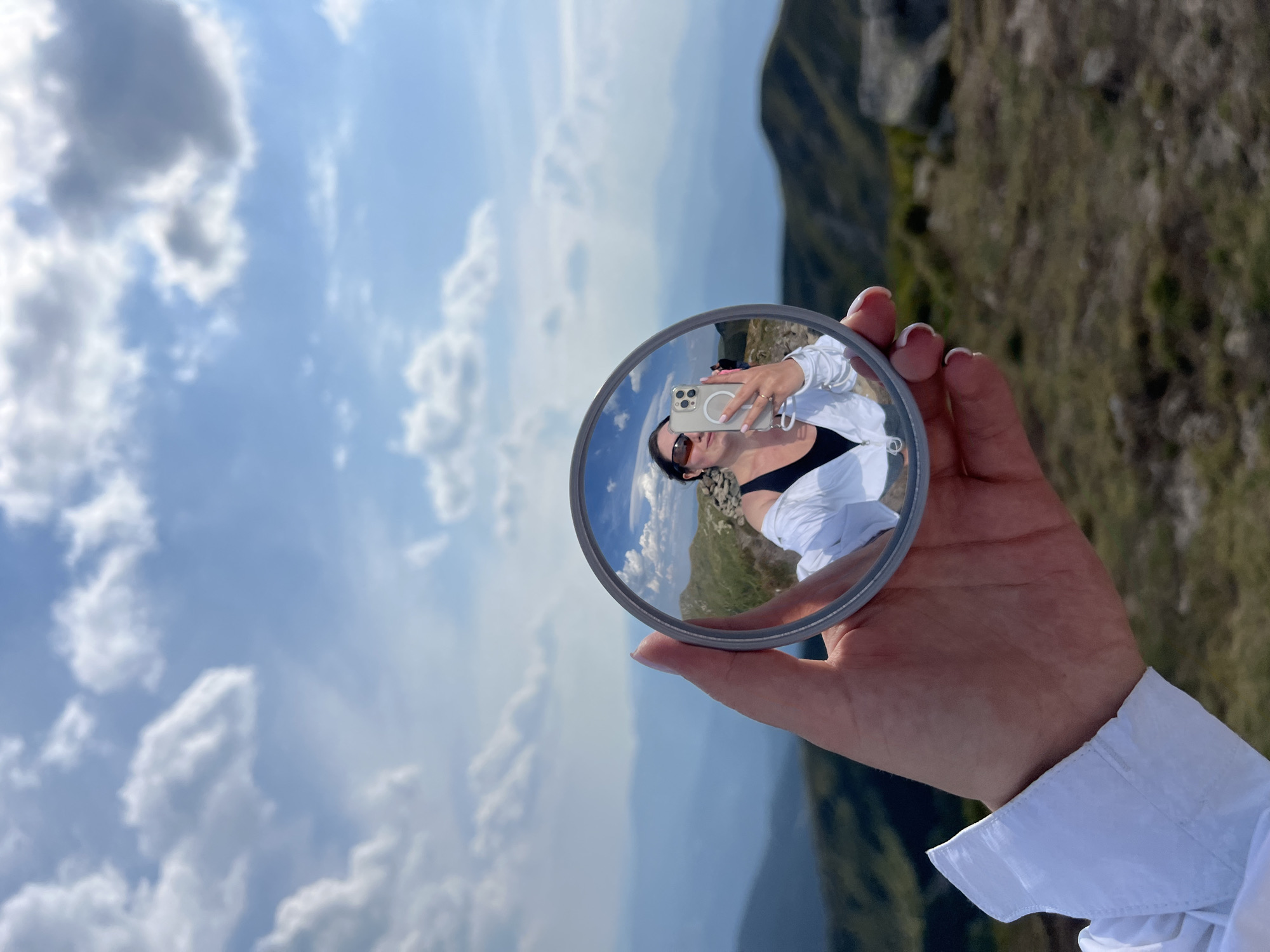 A person's hand, wearing a white shirt, holds up a round mirror, capturing a reflection of themselves and the surrounding mountainous landscape. The person in the reflection is wearing sunglasses and a white jacket over a black top, holding a smartphone to take a selfie. The background is a vast expanse of rolling hills and mountains under a bright blue sky with scattered clouds, suggesting an outdoor setting, likely in a mountainous region. While the exact location in Luhy, Ukraine cannot be confirmed from the image alone, the scenery is consistent with the mountainous terrain found in parts of Ukraine.