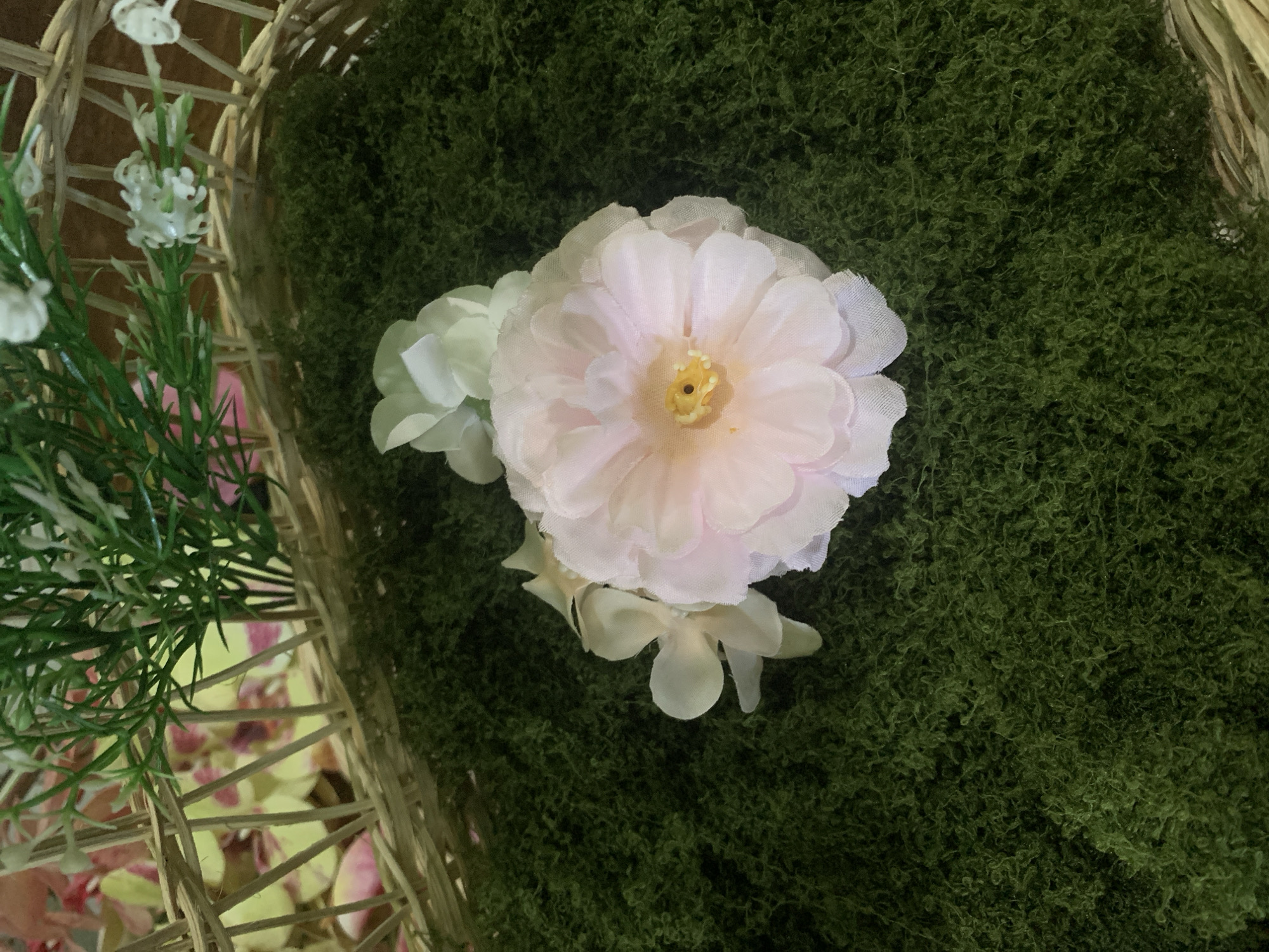 A close-up, top-down shot shows a light pink artificial flower with a yellow center and two smaller light green artificial flowers placed on a bed of green artificial moss. The moss is in a woven basket, and the top portion of the image shows the edge of the basket and some artificial greenery and white flowers spilling over the side. The scene is indoors, likely a decorative arrangement, and not in Kuala Pembuang, Indonesia.