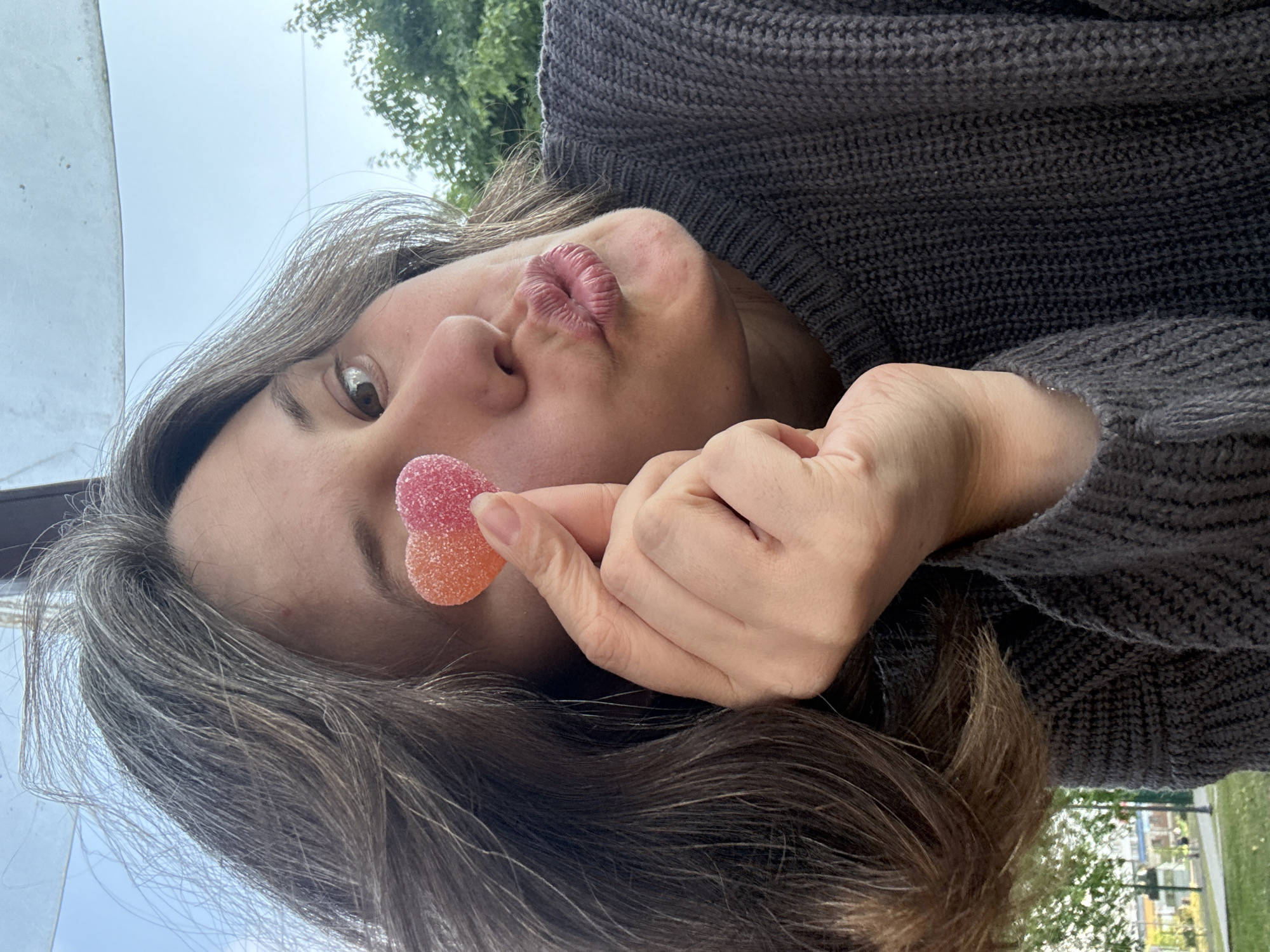 A young woman is taking a selfie outdoors, possibly in the city of Chaikovsky, Russia. She is holding a heart-shaped gummy candy in front of her eye, her lips pursed in a kiss. The woman is wearing a dark grey, knitted sweater. Her hair is long and brown with streaks of grey. The background shows trees and a hint of blue sky.