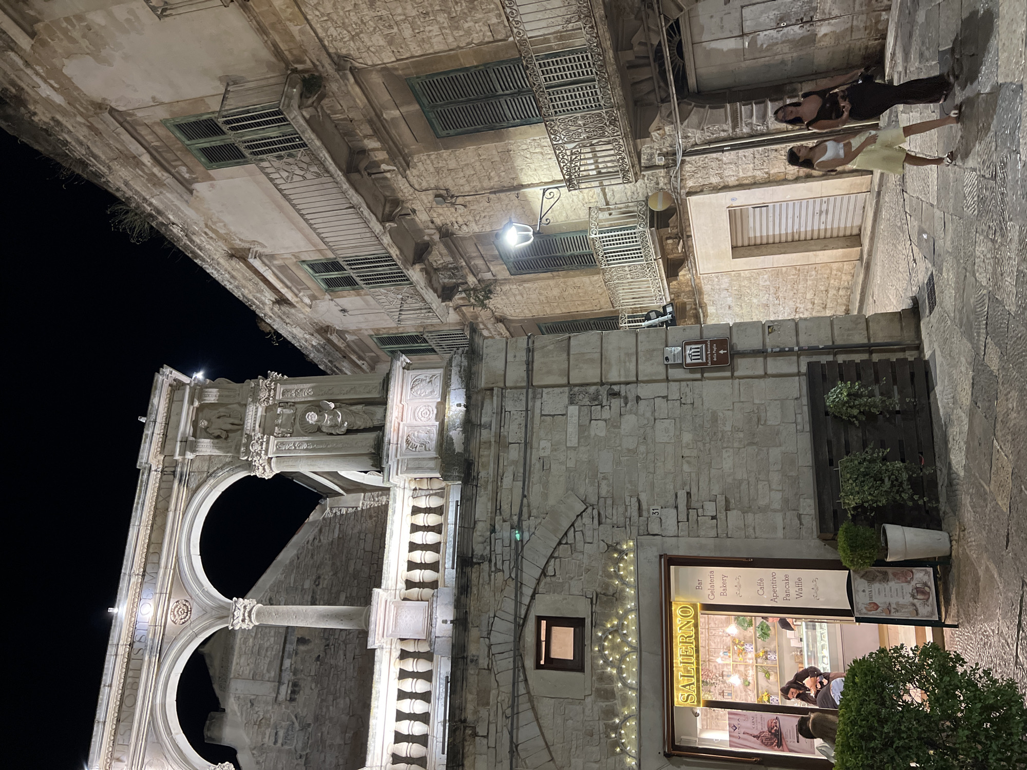 The image shows a street scene at night in the city of Bitonto, Italy. In the foreground, a shop named "SALIERNO" is visible, which appears to be a bar, gelateria, and bakery. A woman is standing inside the shop, and several customers are seated outside. Two women are walking down the street in the middle ground. In the background, an ancient building with arches and statues is illuminated. The street is paved with stone, and there are plants and lighting fixtures visible.