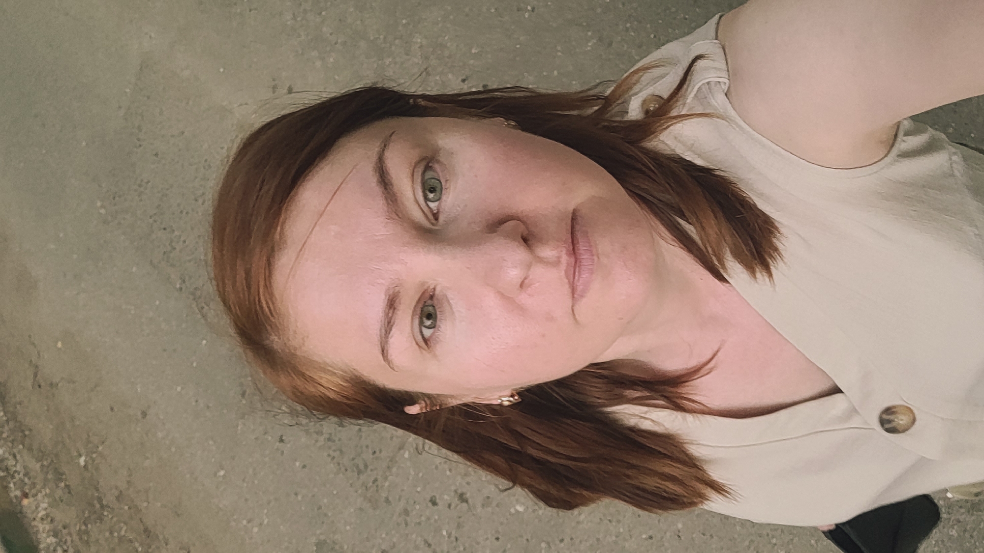 An overhead, eye-level medium shot shows a light-skinned woman with auburn hair and green eyes, looking directly at the camera. She is wearing a light-colored, sleeveless V-neck top with a button detail on the front. The background is a rough, gray concrete surface. The image appears to be a selfie taken outdoors.