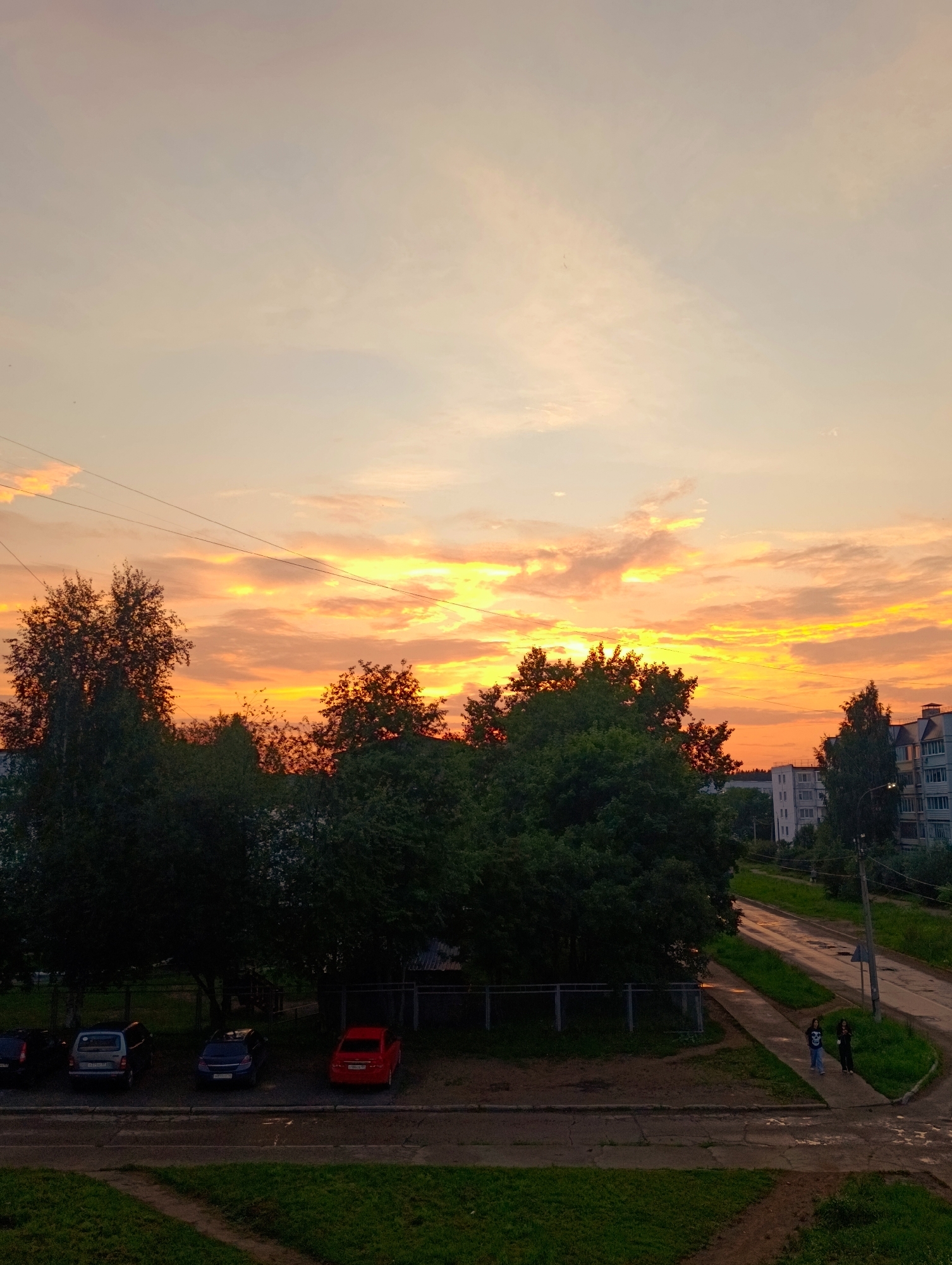 This image depicts a sunset scene in Chaikovsky, Russia, likely during the summer months. The sky is illuminated with vibrant hues of orange and yellow, indicative of a sunset. In the foreground, a row of parked cars, including a red sedan and two SUVs, are visible on a paved surface adjacent to a grassy area. Tall trees with dark green foliage dominate the middle ground, creating a dense line that partially obscures the view of buildings behind them. These buildings appear to be residential apartment blocks, with multi-story structures visible in the distance. A paved road curves through the scene, with two individuals walking along the sidewalk beside it. Overhead, power lines crisscross the sky. The overall impression is a residential urban landscape at dusk, softened by the warm glow of the setting sun.