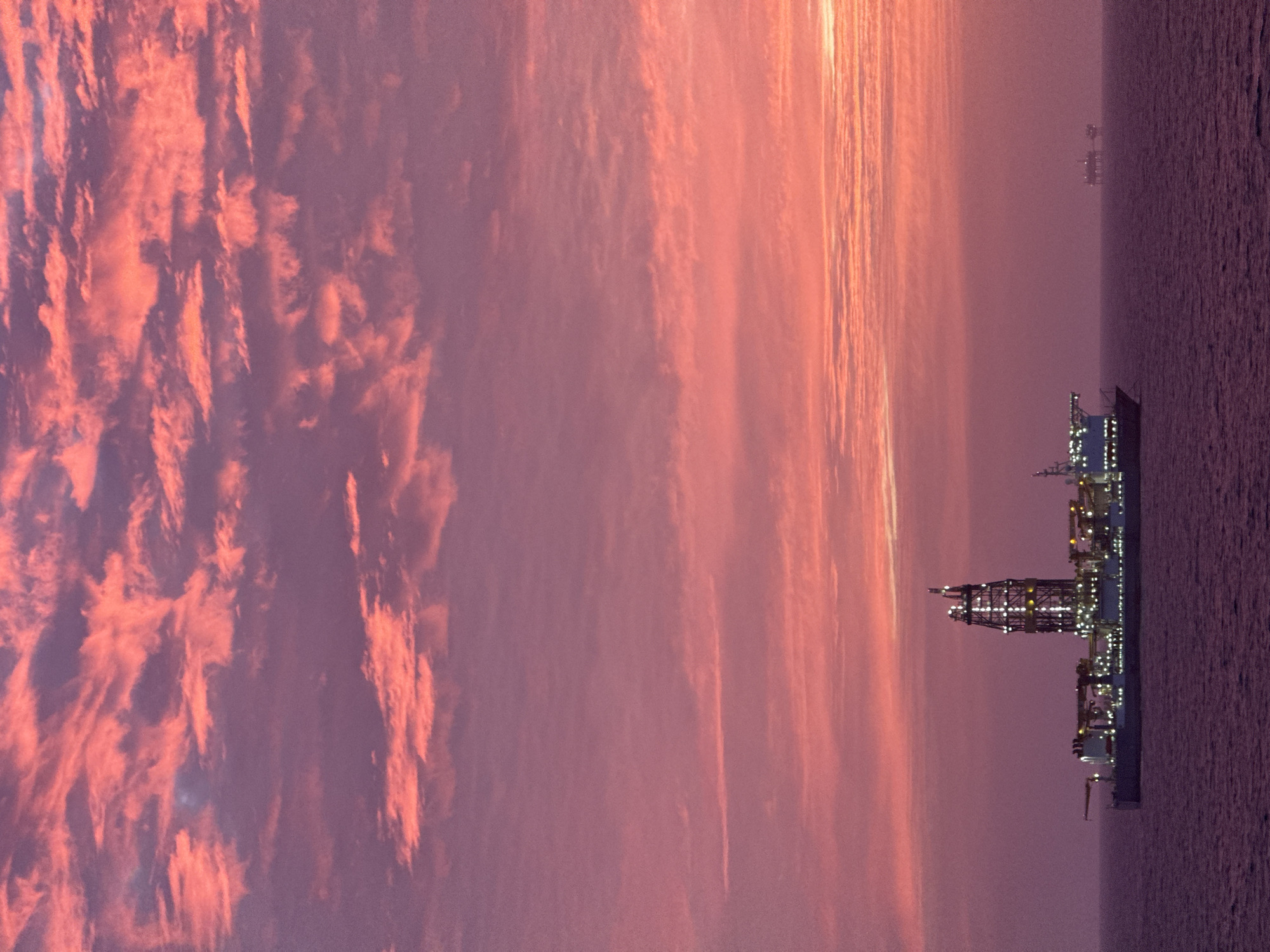 The image depicts an offshore oil platform at dusk. The platform is illuminated with lights, and its structure is visible against the sky and sea. The sky is filled with clouds tinged with pink and orange, suggesting a sunset or sunrise. The sea is calm. Another platform is visible in the distance. The location is Unknown, Unknown. There are no people visible.