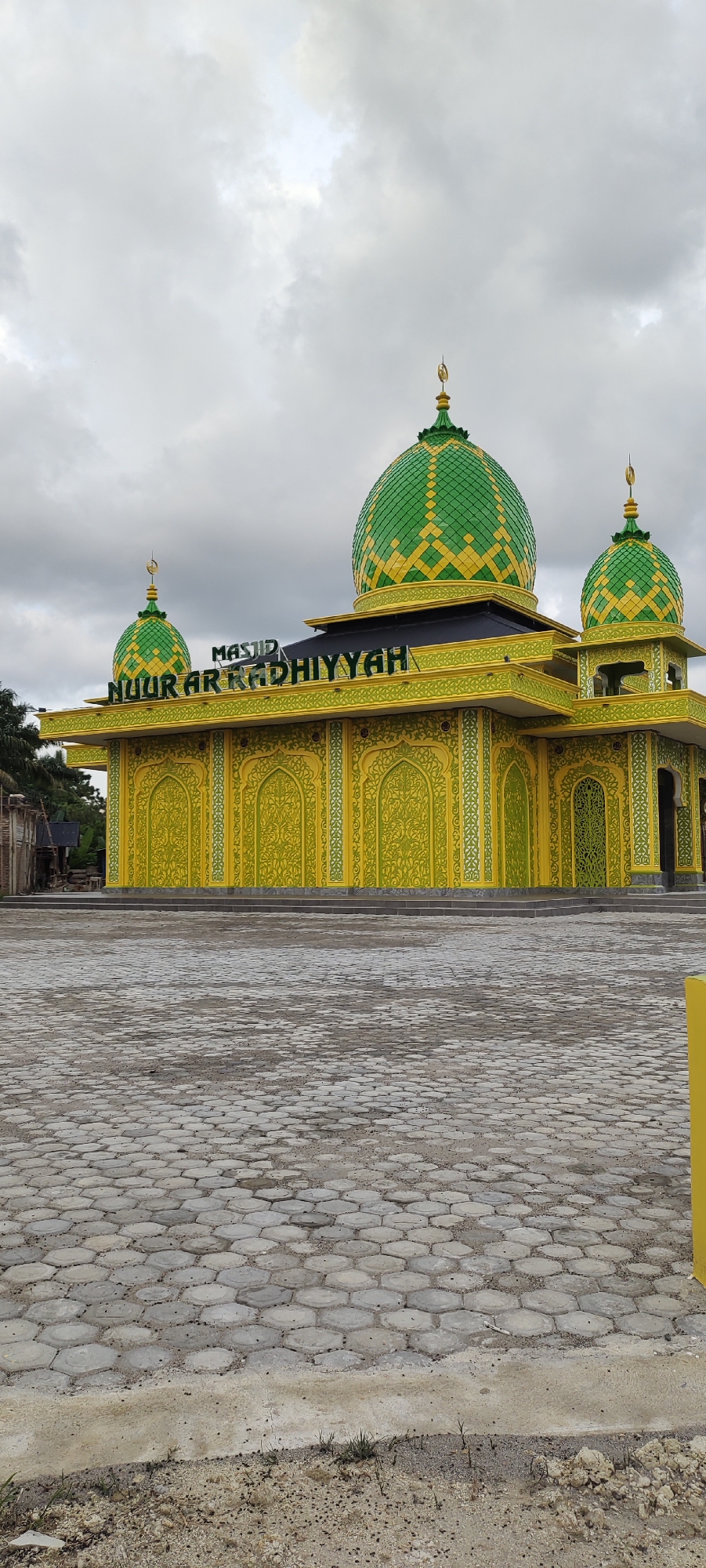 The image shows Masjid Nuur Ar-Radhiyyah, a mosque located in Aluekeujruen, Indonesia. The mosque is painted in bright yellow and green colors. The main dome and two smaller domes are green with yellow diamond patterns. The walls are yellow with green ornamental designs. The name of the mosque is displayed in black letters above the entrance. The ground in front of the mosque is paved with gray hexagonal concrete blocks. The sky is overcast. A yellow pole is visible in the lower right corner. Some trees and a simple wooden structure are visible in the background to the left of the mosque.