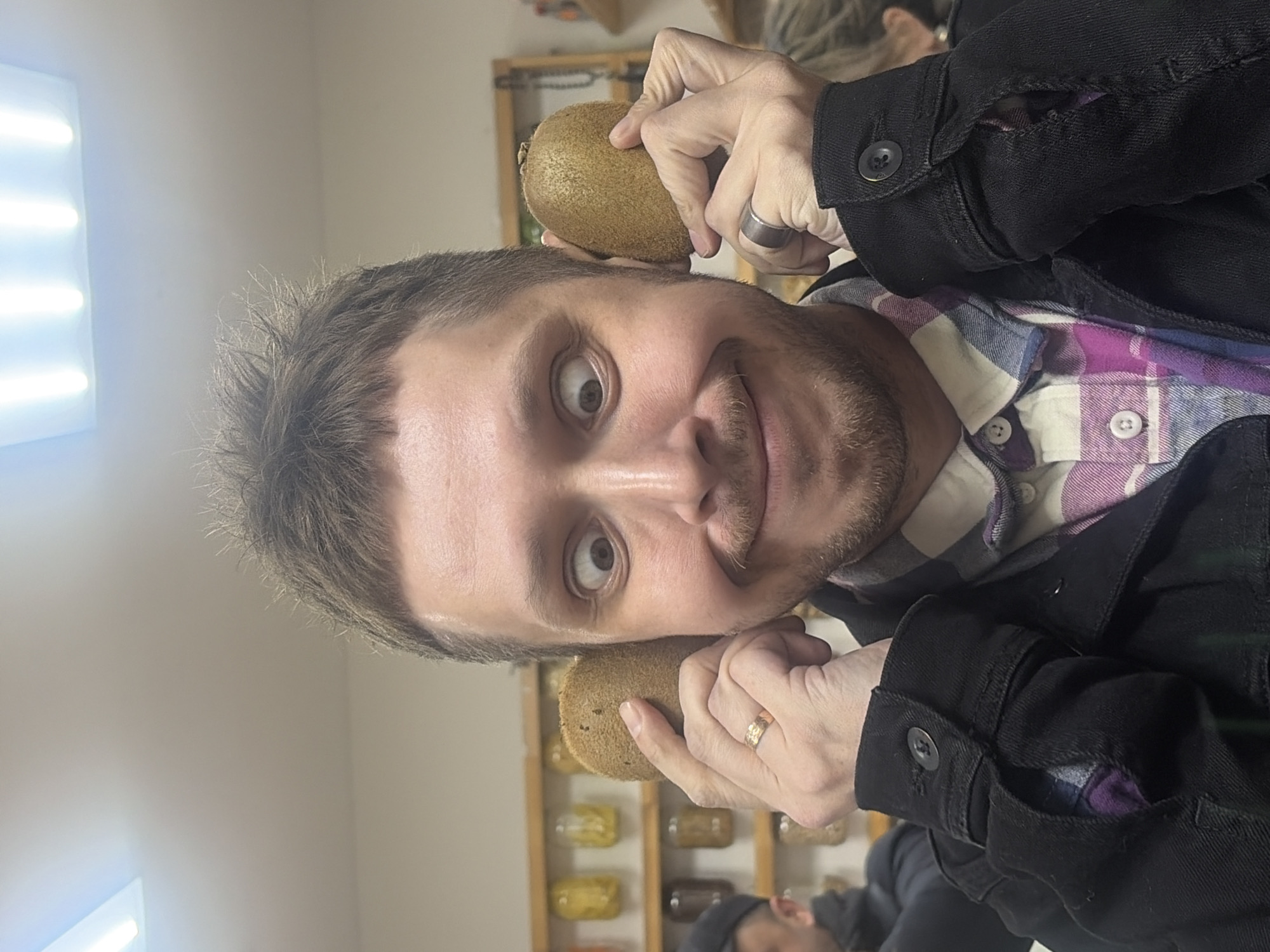 The image features a man’s playful close-up, rotated 90 degrees counter-clockwise. He has light skin, short brown hair, and stubble or a short beard. His eyes are wide open, and he wears a slight smirk, looking directly at the camera. He is wearing a plaid shirt with shades of purple, white, and blue, layered under a black denim-style jacket.
The central activity involves the man holding two kiwis to his ears, one in each hand, as if they are makeshift headphones or earmuffs. A silver band ring is visible on his left hand (upper hand in the rotated image), and a gold band ring is on his right hand (lower hand).
The setting appears to be indoors, likely in a shop or cafe in Saint Petersburg, Russia. In the background, out of focus, there are wooden shelves filled with numerous clear jars containing various food items, possibly dried fruits, nuts, or spices. To the left of the man, a bright, rectangular light fixture with multiple illuminated bars casts a strong artificial light. A second, out-of-focus person is partially visible in the lower right corner of the image. The walls are light-colored. There is no visible text in the image. The time of day cannot be determined due to the artificial lighting.