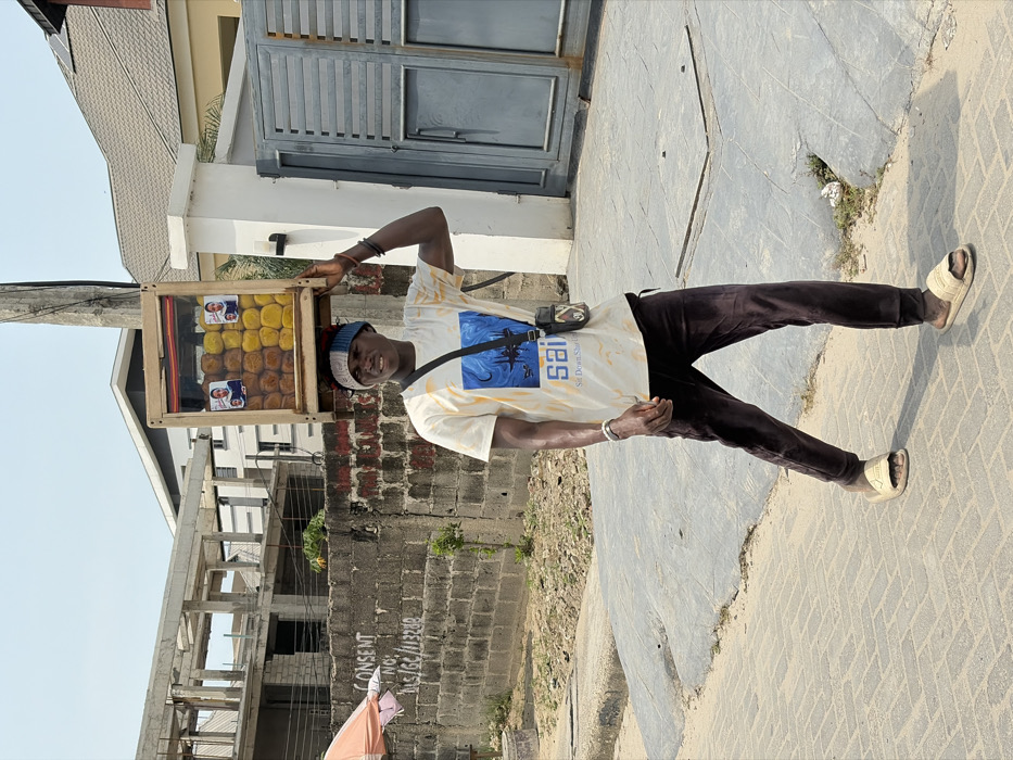 A young adult male walks on a paved sidewalk in Igboefon, Nigeria, under a clear sky. He wears a white t-shirt with a blue graphic featuring the text "sai" and "Sit Down Shut Up," dark brown velvet-like pants, and light beige slide sandals. A multi-colored knitted headband adorns his head. He carries a small dark shoulder bag across his body and a silver-colored bracelet on his left wrist. Balanced on his head and supported by his left hand is a rectangular wooden display case with a clear front panel, containing rows of brown and yellow spherical food items. Two small laminated portraits are affixed to the front of the display case. The individual is smiling and looking towards the viewer. In the background, to the left, a concrete block wall with the legible marking "CONSENT NO: DIS/GC/11328" and an unfinished concrete building structure are visible. To the right, a grey metal gate, a concrete pillar, and a modern building with a grey roof are present. A utility pole stands near the gate. The sidewalk is constructed from light-colored rectangular pavers, with a dirt shoulder alongside.