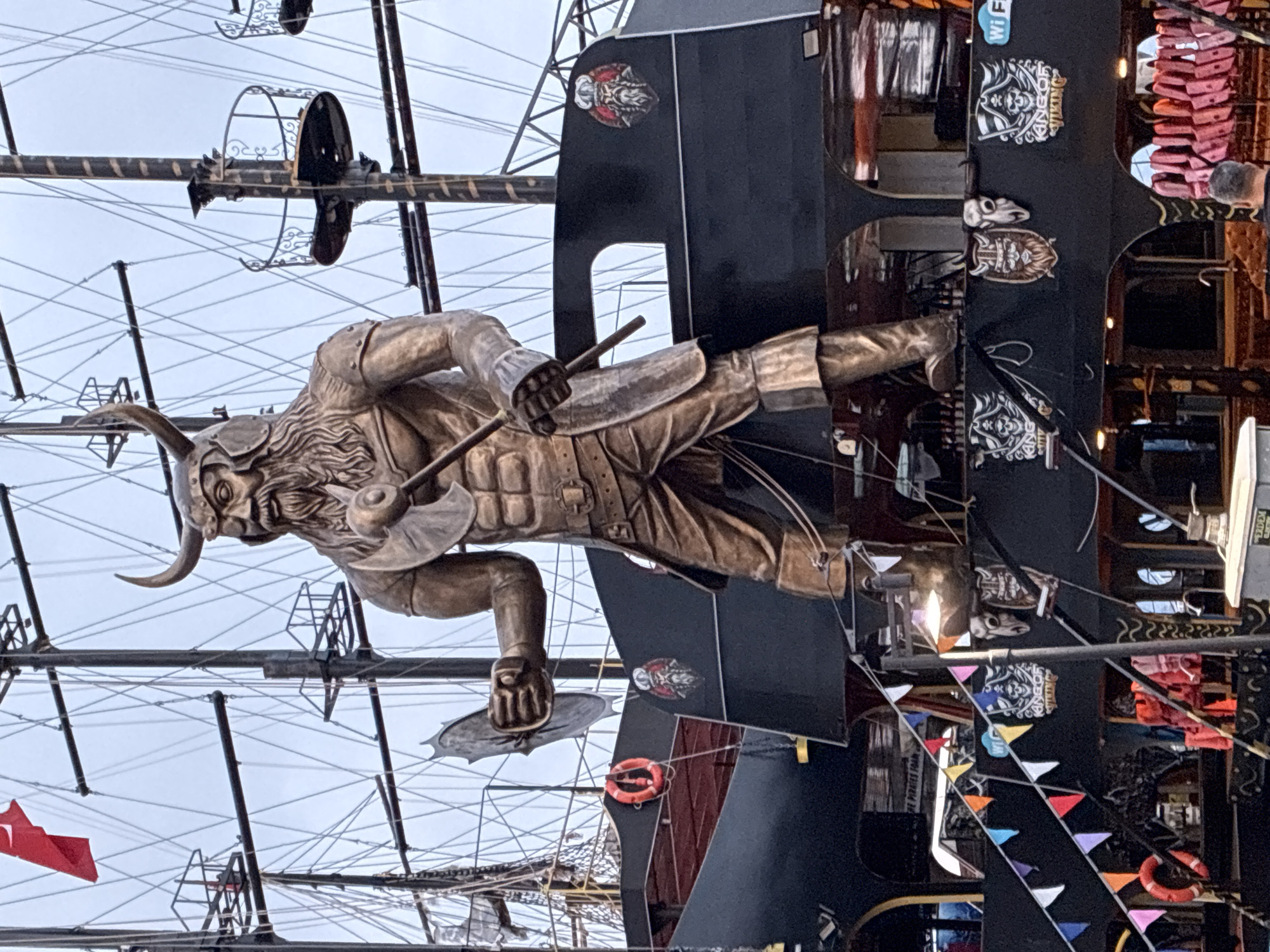 This image captures a large, elaborately decorated ship, likely a pirate or Viking-themed tourist vessel, under an overcast sky in Alanya, Turkey.
The central subject is a prominent, bronze-colored statue of a Viking warrior or pirate, complete with a horned helmet and a flowing beard. He holds a staff or club in his right hand and a round shield in his left, standing in a powerful, dynamic pose on the side of the ship, serving as an ornate figurehead.
The ship itself features a dark (possibly black) hull or superstructure. Behind the statue, numerous masts and intricate rigging create a complex web against the pale, cloudy sky, indicating a multi-masted sailing ship. The ship's side is decorated with colorful triangular pennants strung along its length. Several stylized logos depicting Viking or pirate-like figures are affixed to the ship's exterior, some clearly displaying the text "KING OF VIKING." A red lifebuoy is visible on the ship's side, along with a "Wi Fi" symbol. Glimpses of the ship's interior or upper deck show curtains and what might be seating, suggesting it functions as a tour boat, restaurant, or bar. A person's back, wearing a dark top, is partially visible on the right.
The scene takes place during the day, under an overcast sky, suggesting mild weather. The ship appears to be stationary, likely docked or anchored. The overall ambiance is one of adventure and entertainment, characteristic of a tourist attraction in a coastal resort area.