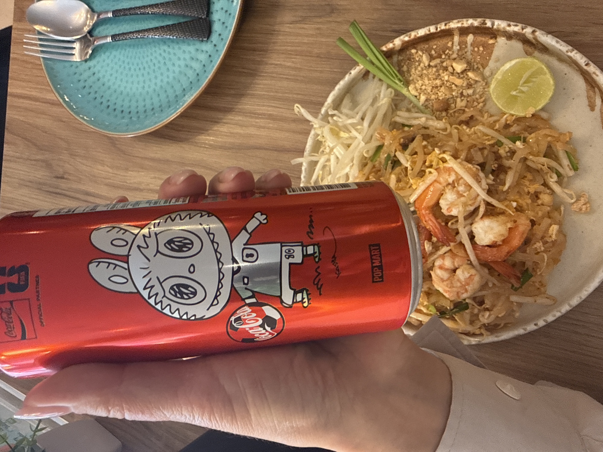 The image depicts a close-up, eye-level view of a person's hand holding a red Coca-Cola can with a cartoon character design. The hand, adorned with long, manicured fingernails, grips the can from the left side of the frame. The can features a black and white drawing of a character with large eyes, pointed ears, and wearing a soccer jersey. Text on the can includes "Coca-Cola" and "POP MART".
In the background, a plate of Pad Thai, a popular Thai noodle dish, is visible. The dish contains noodles, shrimp, bean sprouts, and is garnished with chopped peanuts, green onions, and a lime wedge. To the right of the plate, a turquoise textured plate holds a fork and spoon. The setting appears to be a restaurant with a wooden table surface. The image does not provide any specific visual cues that would definitively place the scene in Pattaya City, Thailand; it is a generic restaurant setting.