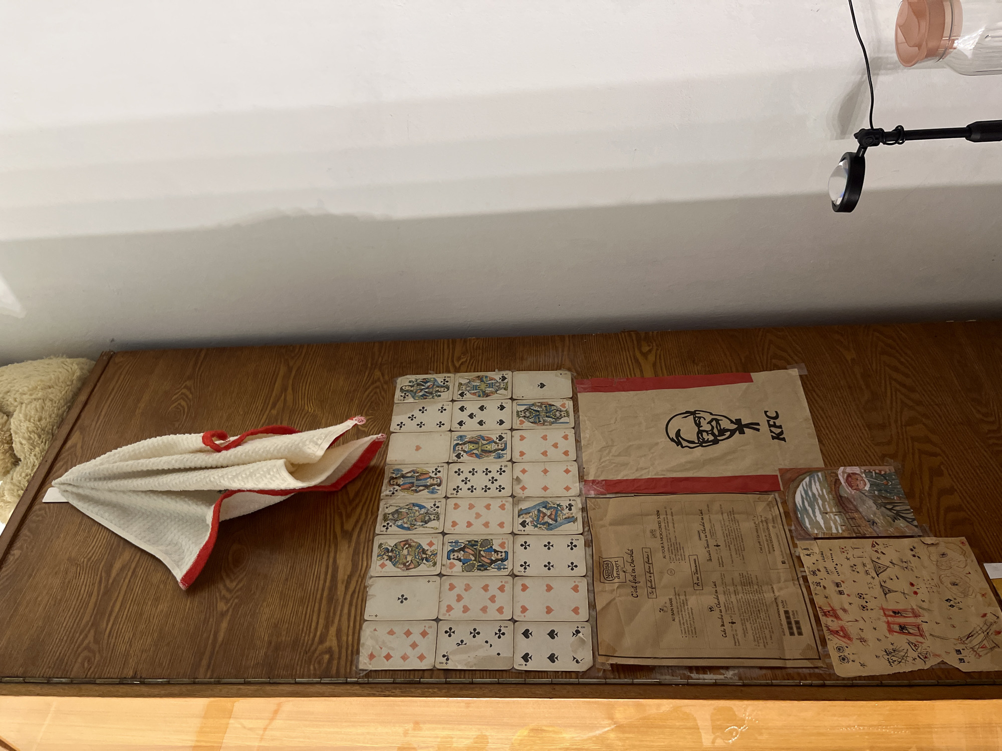 A high-angle shot depicts a wooden table cluttered with various items. On the left, a folded white towel with a red border sits next to a pile of playing cards arranged in rows and columns. Adjacent to the cards, there is a KFC paper bag with the KFC logo prominently displayed. Next to the bag there is a recipe for Nestle dessert in French. Finally, there are two pieces of paper with drawings. The location is not identifiable as Perm, Russia based on the items visible in the photo.