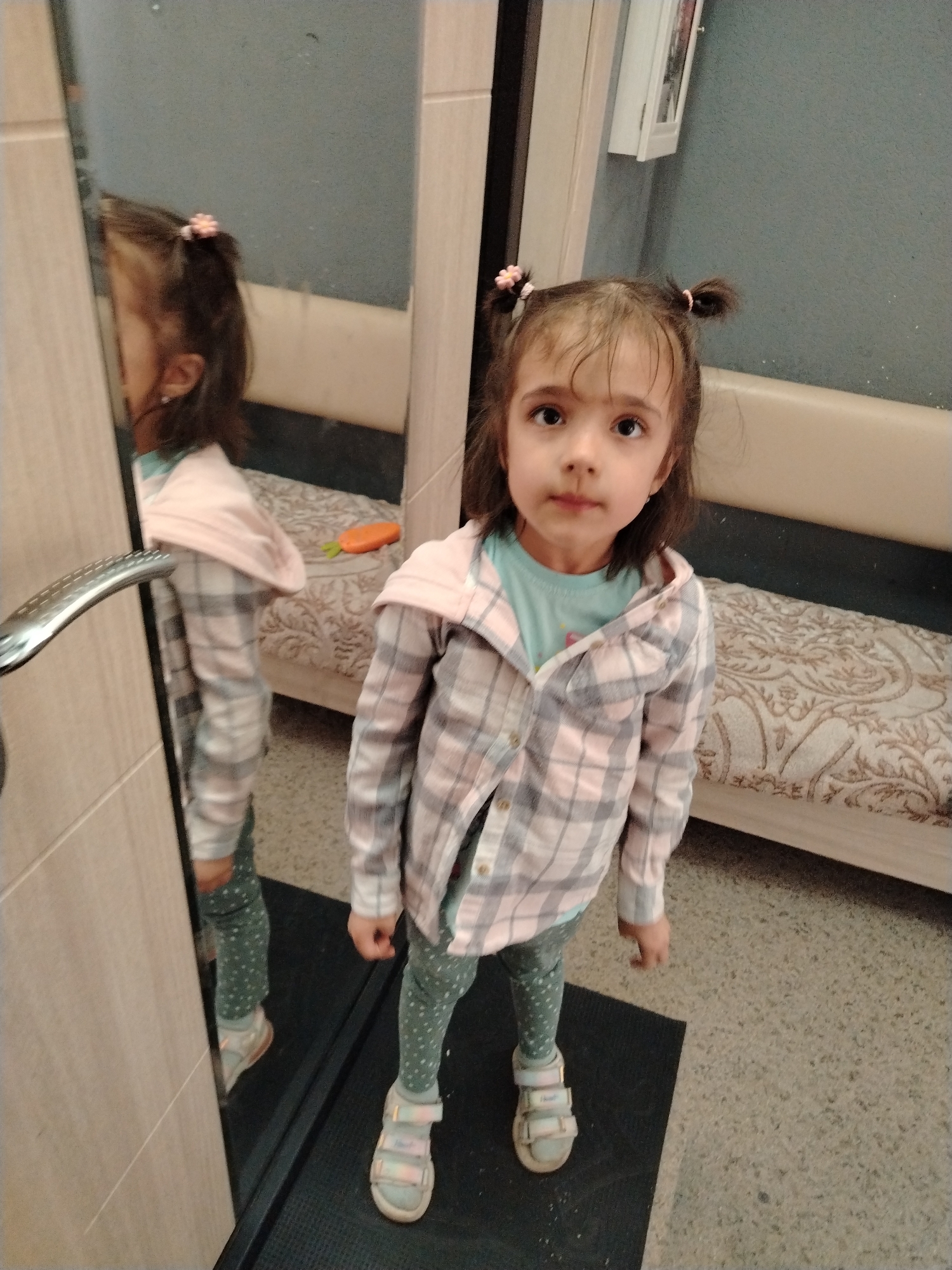 A young girl stands on a black mat in front of a mirror, looking directly at the camera. She has brown hair styled in two small pigtails adorned with pink flower clips. She wears a light blue t-shirt, a pink and gray plaid open shirt, patterned leggings, and sandals. The mirror reflects her image, along with a portion of a sofa or bench covered in a patterned beige fabric. An orange toy or item sits on the sofa in the reflection. The background appears to be a room with gray walls, and a framed picture hangs on the wall above the sofa. Based on visual cues, the location is Noviy, Russia.