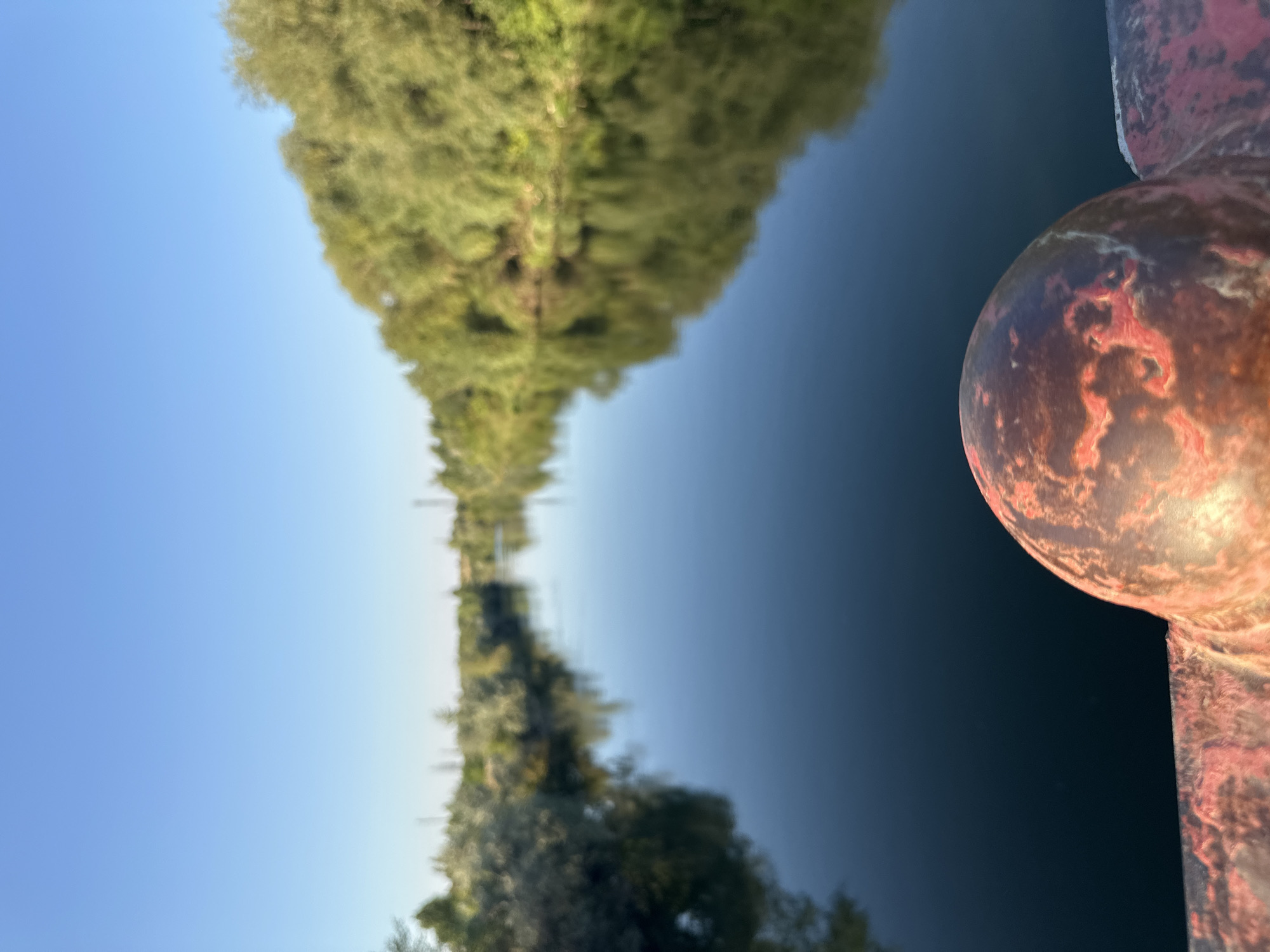 The image shows a calm river with trees lining its banks. The sky is clear and blue. In the foreground, there is a blurred reddish-brown railing. The scene appears to be a peaceful natural landscape. The location is identified as Energetik, Russia. There are no people or animals visible in the image. The focus is on the tranquil river and the surrounding vegetation. The time of day appears to be daytime, given the bright sky and sunlight.