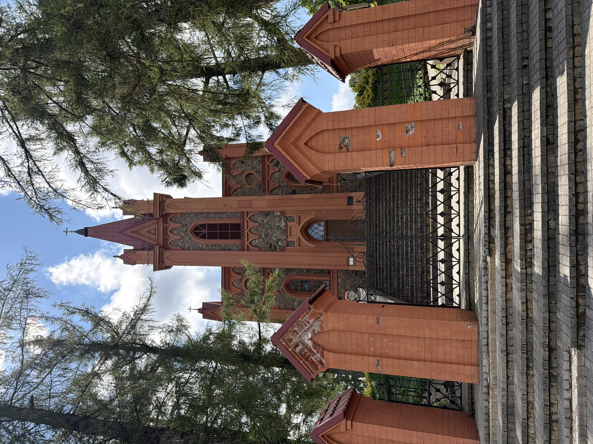 The image captures an exterior view of the Church of the Holy Trinity in Dūkštos, Lithuania, on a bright, partly cloudy day. The church, constructed primarily of red brick and stone, features a prominent central tower with a steeple and red roof. Gothic-style arched windows and intricate brickwork are visible on its facade. In the foreground, a wide set of stone stairs leads up to a decorative black metal gate, which is flanked by red brick pillars topped with small, pointed roofs. Mature evergreen trees with sparse foliage frame the upper portion of the image, and the sky is dotted with white clouds. No people or animals are discernibly present.