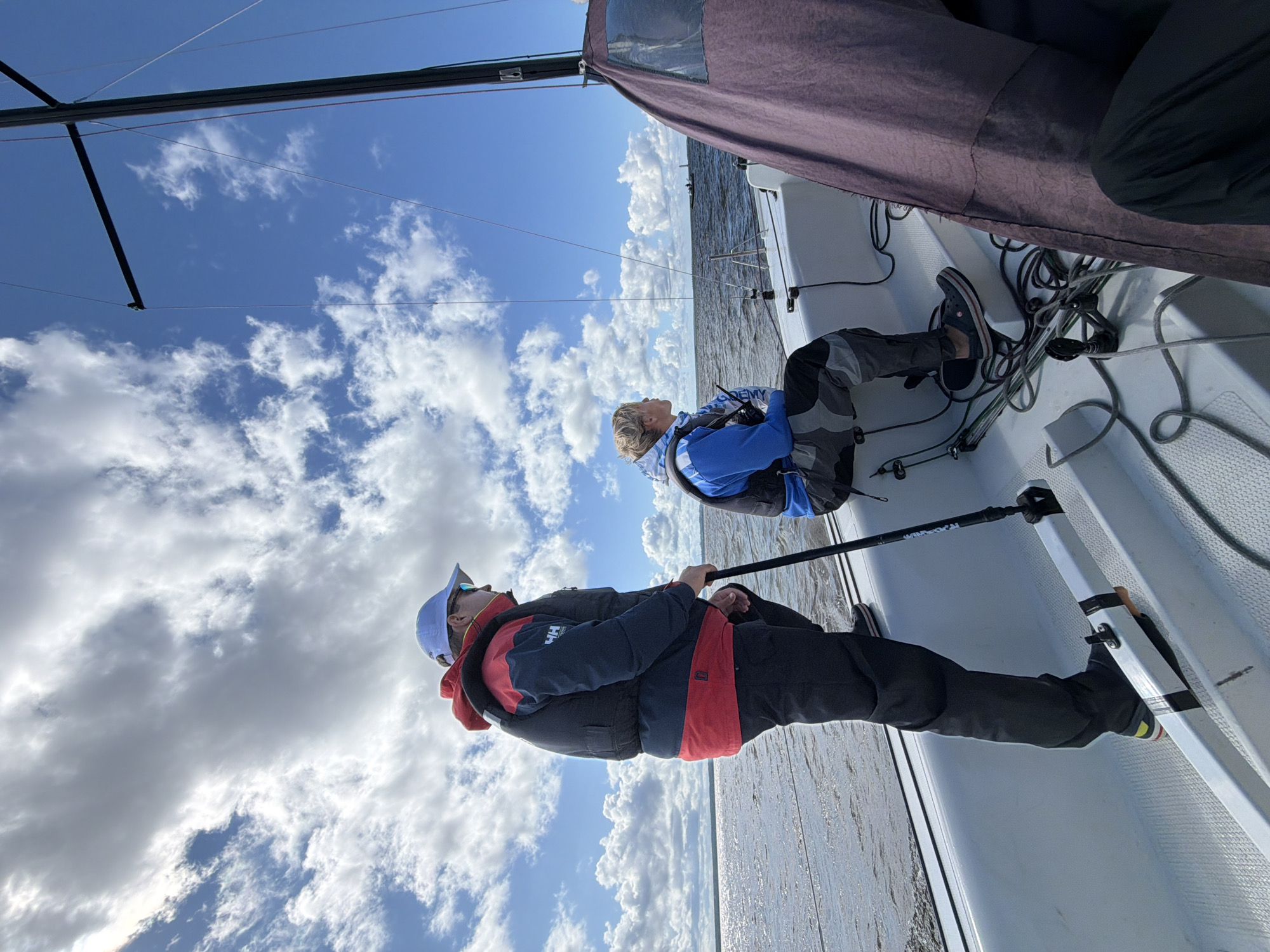 Two individuals are on a white sailboat, looking out at the water under a blue sky with scattered clouds. The person in the foreground, wearing a red and dark blue jacket and a white cap, is standing and holding a black pole. To their right, a second person, wearing a blue jacket, is seated. The sailboat's mast and some rigging are visible. The water appears choppy, suggesting a breeze. In the distance, a small structure, possibly a lighthouse, is visible on the horizon. The scene appears to be in or near Kronstadt, Russia, a port city known for its maritime history and fortifications.