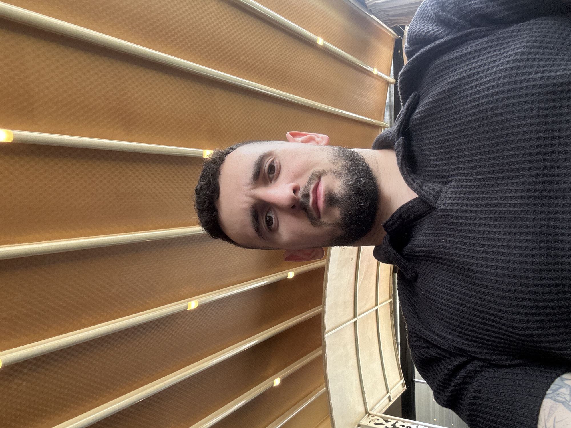 A close-up selfie features a man with short, dark hair and a full beard, partially visible against a background consisting of tan, horizontal, textured panels divided by what appear to be illuminated bars. The man is wearing a black waffle-knit hoodie or sweater. The photo seems to have been taken from a low angle, looking upwards.
Without additional information, pinpointing the specific location in Varna, Bulgaria, is not possible.