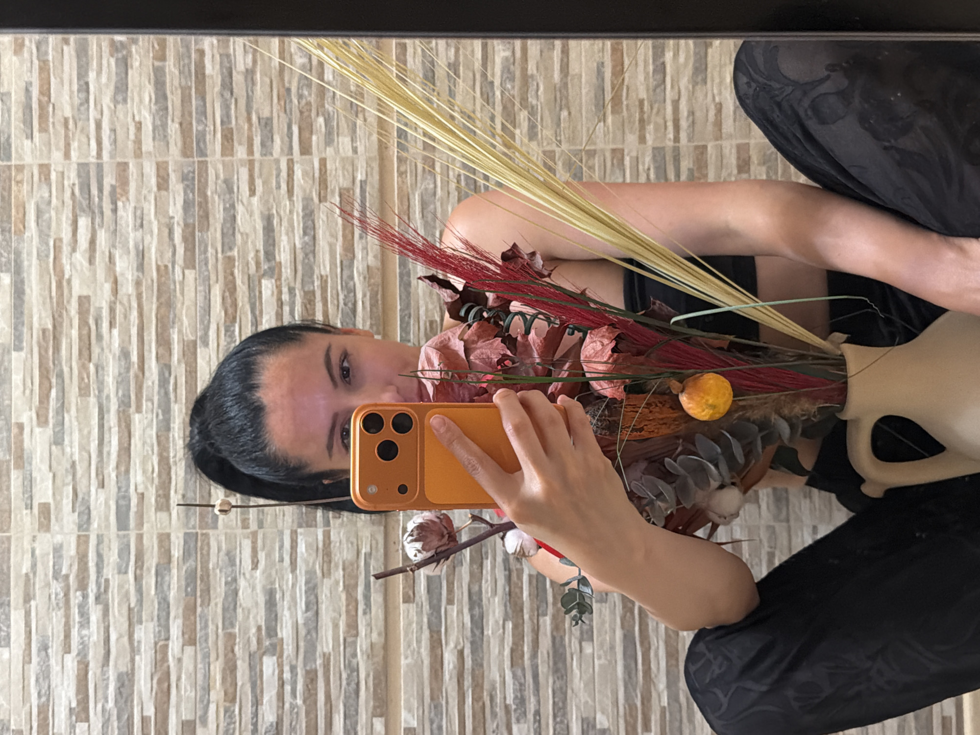 The image shows a woman holding a smartphone in front of her face, taking a selfie in what appears to be a bathroom with textured, light brown and grey tiled walls. She is wearing a black top and black patterned pants. She is sitting with her legs spread apart. In front of her, there is a vase with dried floral arrangements. The bouquet includes red, yellow, and brown dried grasses and leaves, as well as a small orange decorative item and a sprig of what looks like cotton. The woman's left hand is visible, holding the orange-colored smartphone. The overall impression is a casual and intimate moment captured by the woman herself. There is no discernible information within the image that would indicate a specific location in the city of Pleven, Bulgaria.