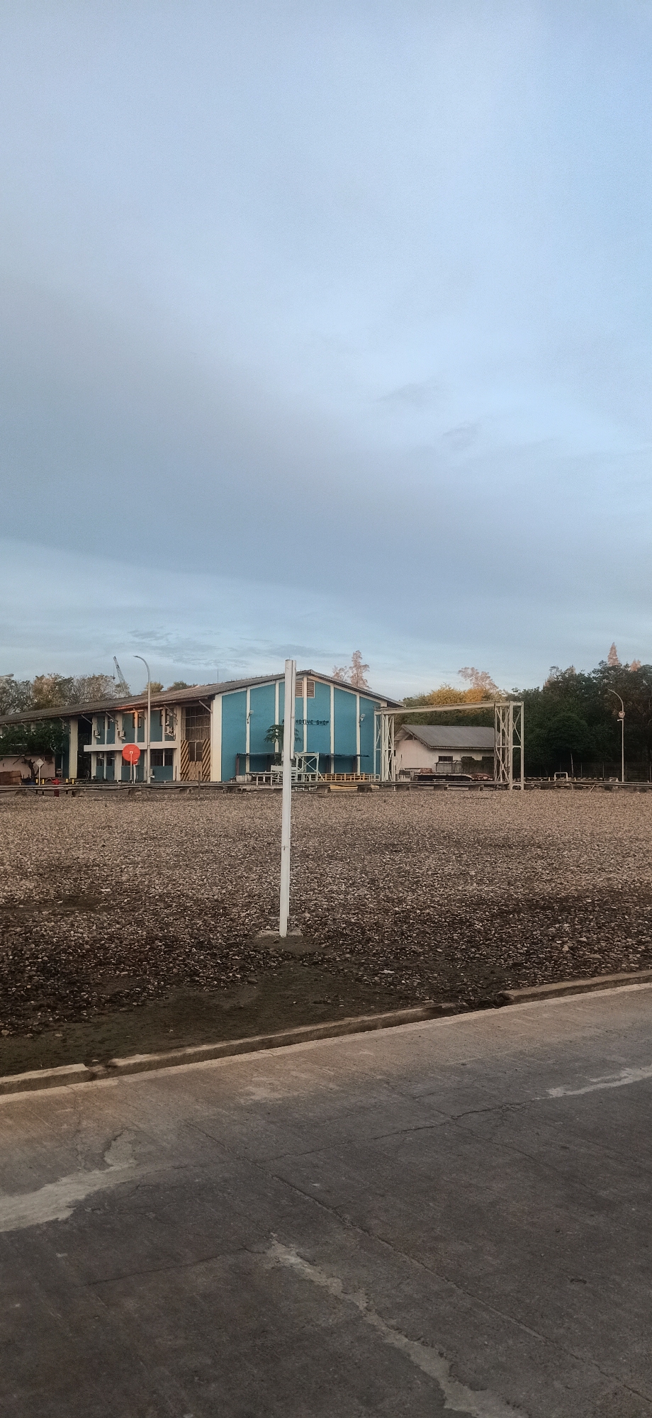 The image shows a long, blue building with the words "AUTOMOTIVE SHOP" in white text visible on its side. To the left of the blue building is a smaller, white building. In front of the buildings is a large, open area covered in gravel. A paved road runs along the bottom of the image, with a concrete curb separating it from the gravel area. A white pole stands in the foreground. Trees and foliage are visible behind the buildings, and the sky is overcast. The scene appears to be in an industrial or commercial area in Palohlada, Indonesia. There are no people visible in the image.