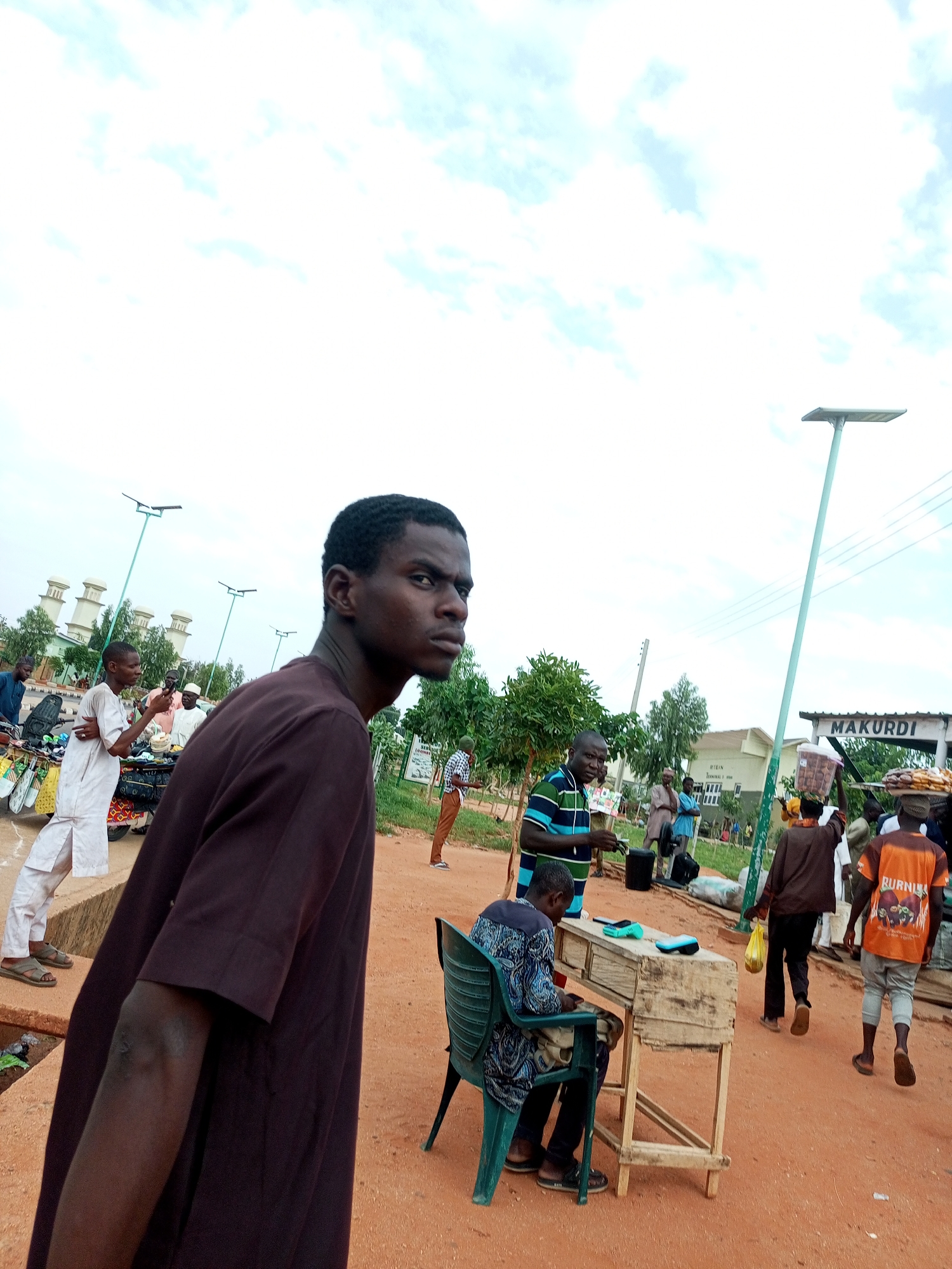 This is a medium-shot, eye-level, outdoor photograph taken in what appears to be a market or public gathering area in Gombe, Nigeria. The foreground is dominated by a young man in a dark brown tunic, facing away from the camera and looking to his right. His expression is serious, and his head is slightly turned.
In the mid-ground, several individuals are engaged in various activities. To the left, a man in a white traditional garment stands by a cart filled with goods, possibly merchandise. Further to the right, a man in a striped t-shirt is interacting with another person, and another man in a light brown tunic and a white cap is visible. A woman is seen carrying a large container of goods on her head. A street vendor's stall, constructed from wood, is positioned in the center-right, with a person seated behind it.
The background shows a street lamp with a solar panel, a sign that reads "MAKURDI," and buildings in the distance, including some with distinctive architecture, possibly religious or governmental. Trees and greenery are scattered throughout the scene. The ground is covered in reddish-brown earth, typical of the region. The sky is overcast with a mix of white and light blue clouds. The overall atmosphere suggests a bustling commercial or social hub.
