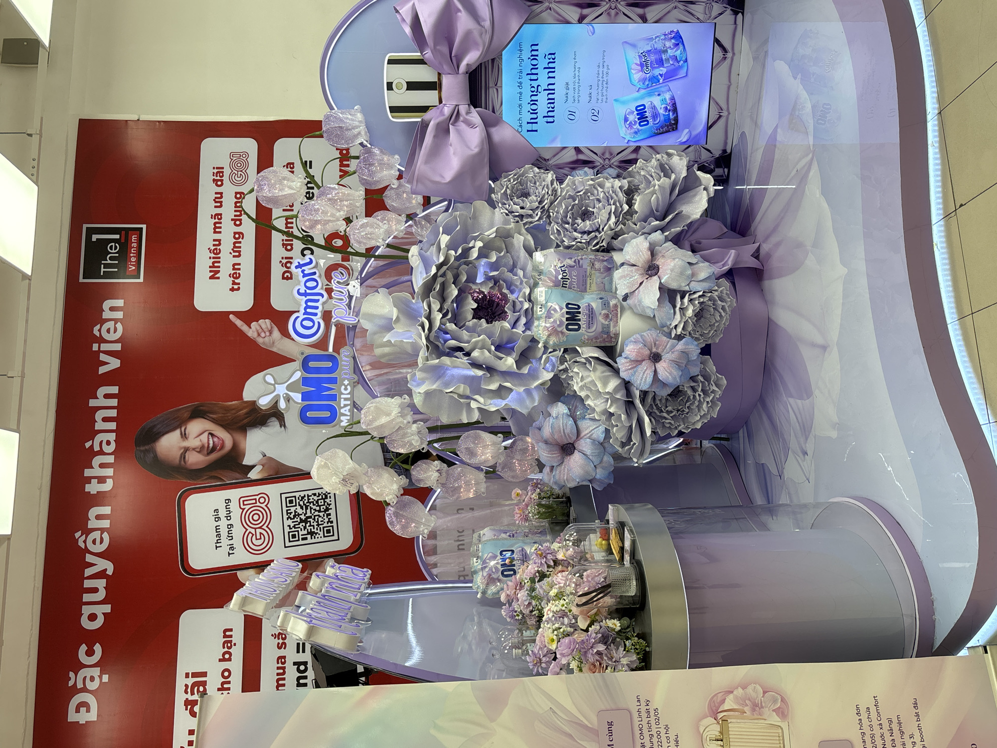 A marketing display for OMO and Comfort Pure laundry products is shown indoors with a red backdrop and a woman in the background. The display features various flowers and products arranged in a tiered design. A QR code is visible on the backdrop. The location is in Đà Nẵng, Vietnam, based on the text on the display. The display includes information about the products' fragrance and benefits.