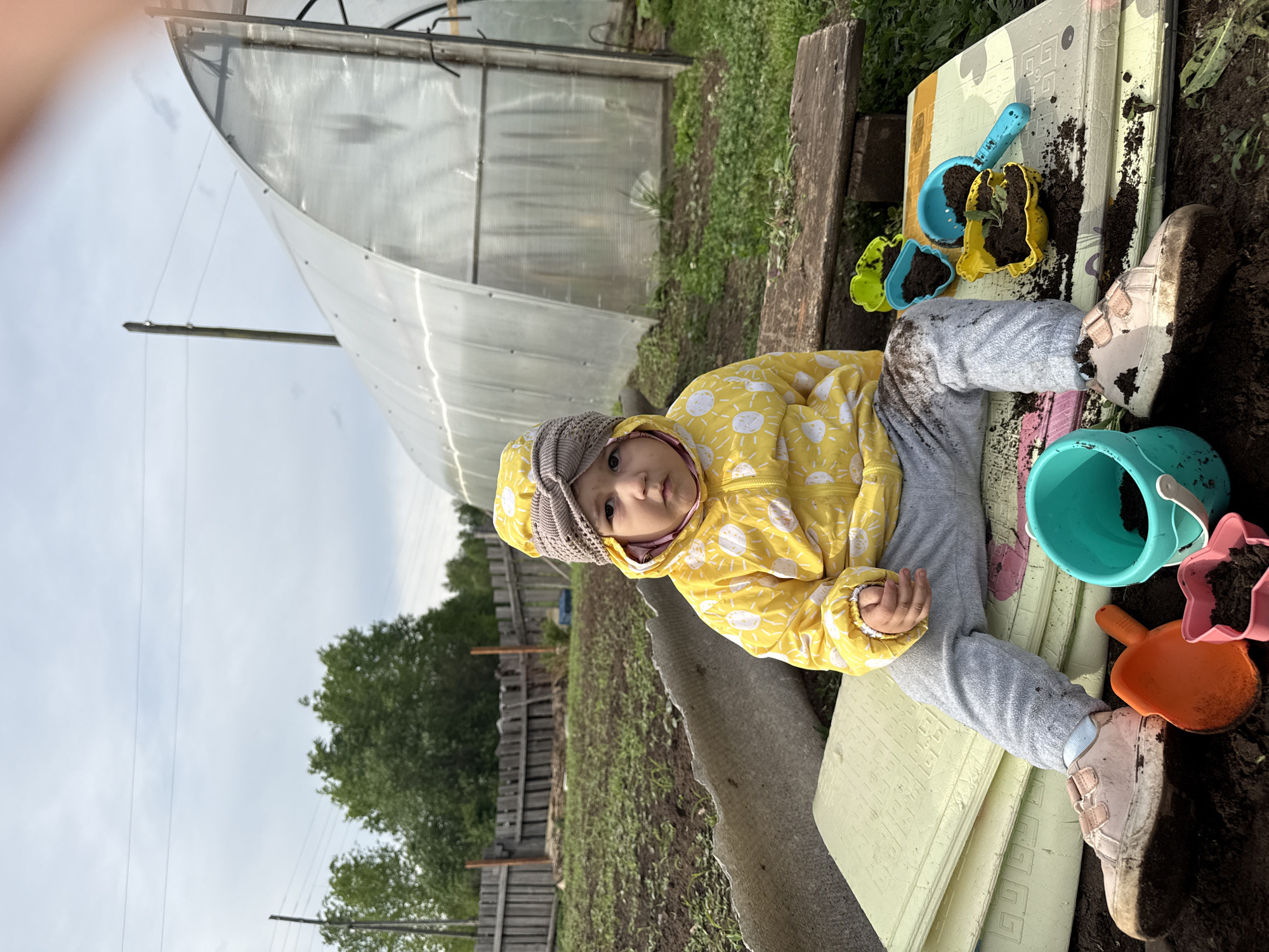 A child wearing a yellow jacket and gray pants sits on a light green mat with various colorful plastic toys, including a blue bucket, orange shovel, and several molds filled with soil. The child appears to be playing with dirt. The scene is outdoors in a garden setting with a greenhouse visible in the background. The garden has rows of plants and a wooden fence. Based on the location provided, this scene is in Уральское, Russia.