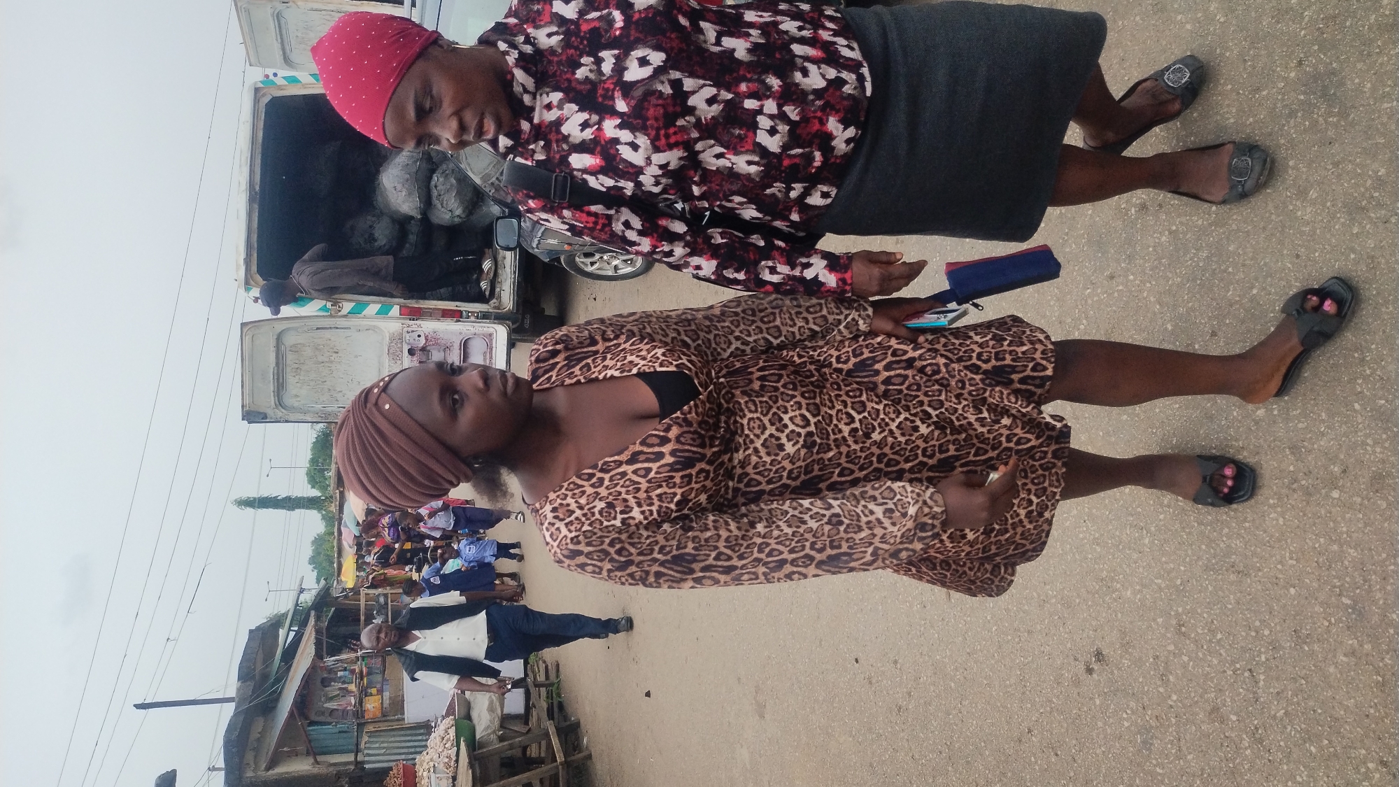 Two women stand on a paved surface next to a vehicle with its back doors open and items in the back. One woman wears a red headscarf and a red, white, and black patterned shirt with a dark skirt and dark-colored flats. The other woman wears a brown headscarf and a brown animal print dress and black sandals. The woman in animal print dress holds a cell phone and something else in her hands. In the background, a market scene unfolds with stalls and people. The location is in Jos, Nigeria.