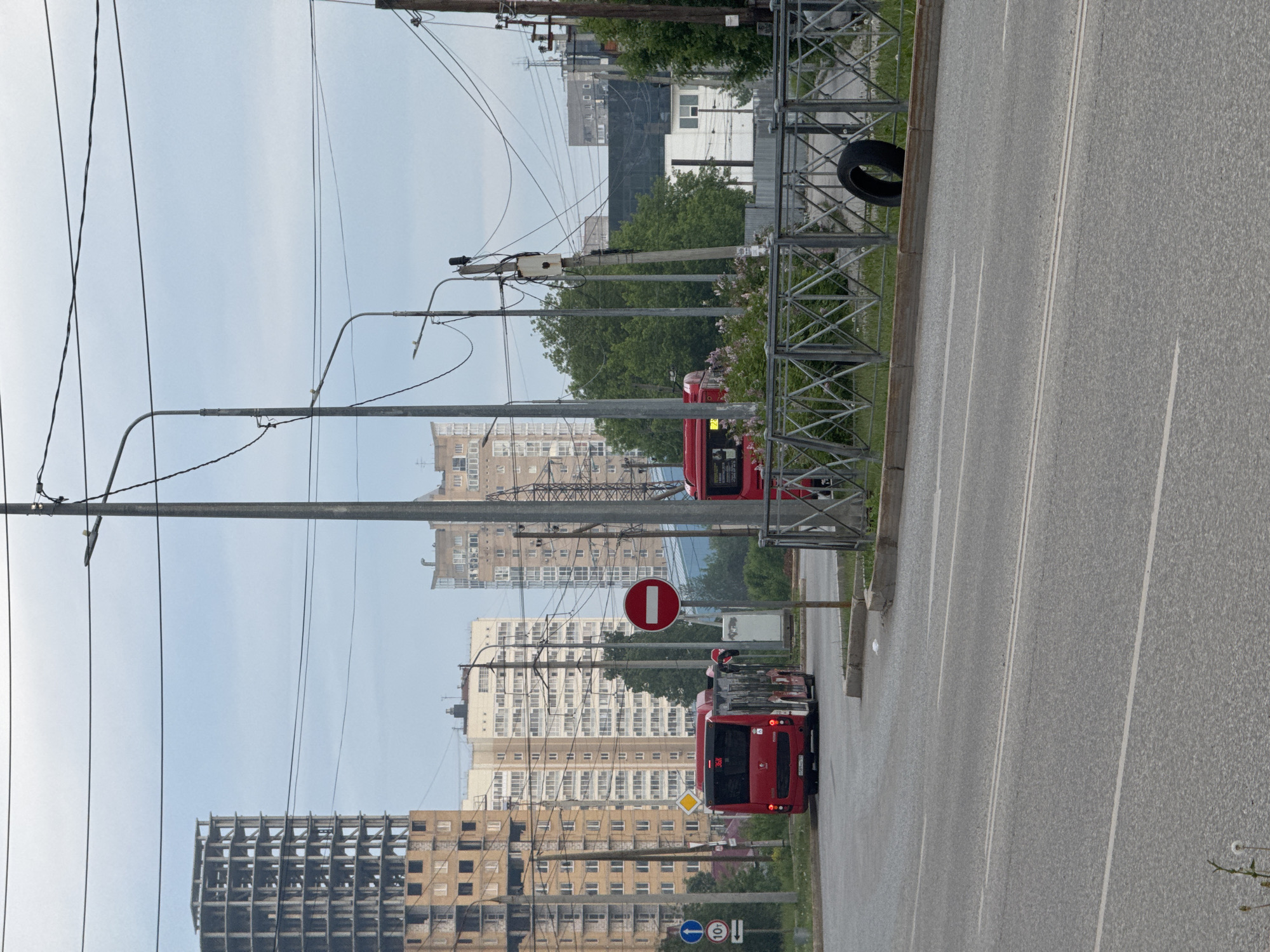 The image depicts a street scene in Perm, Russia, with a focus on urban infrastructure and transportation. A multi-lane asphalt road stretches into the distance, marked by white lane dividers. A protective metal barrier runs along the roadside, with a tire oddly placed against it. Overhead, a complex network of electrical wires and utility poles crisscrosses the sky. In the background, a mix of residential and commercial buildings of varying heights dominate the skyline, including one building under construction. Two red buses are visible on the road. Traffic signs are present, including a 'Do Not Enter' sign. The sky is overcast.