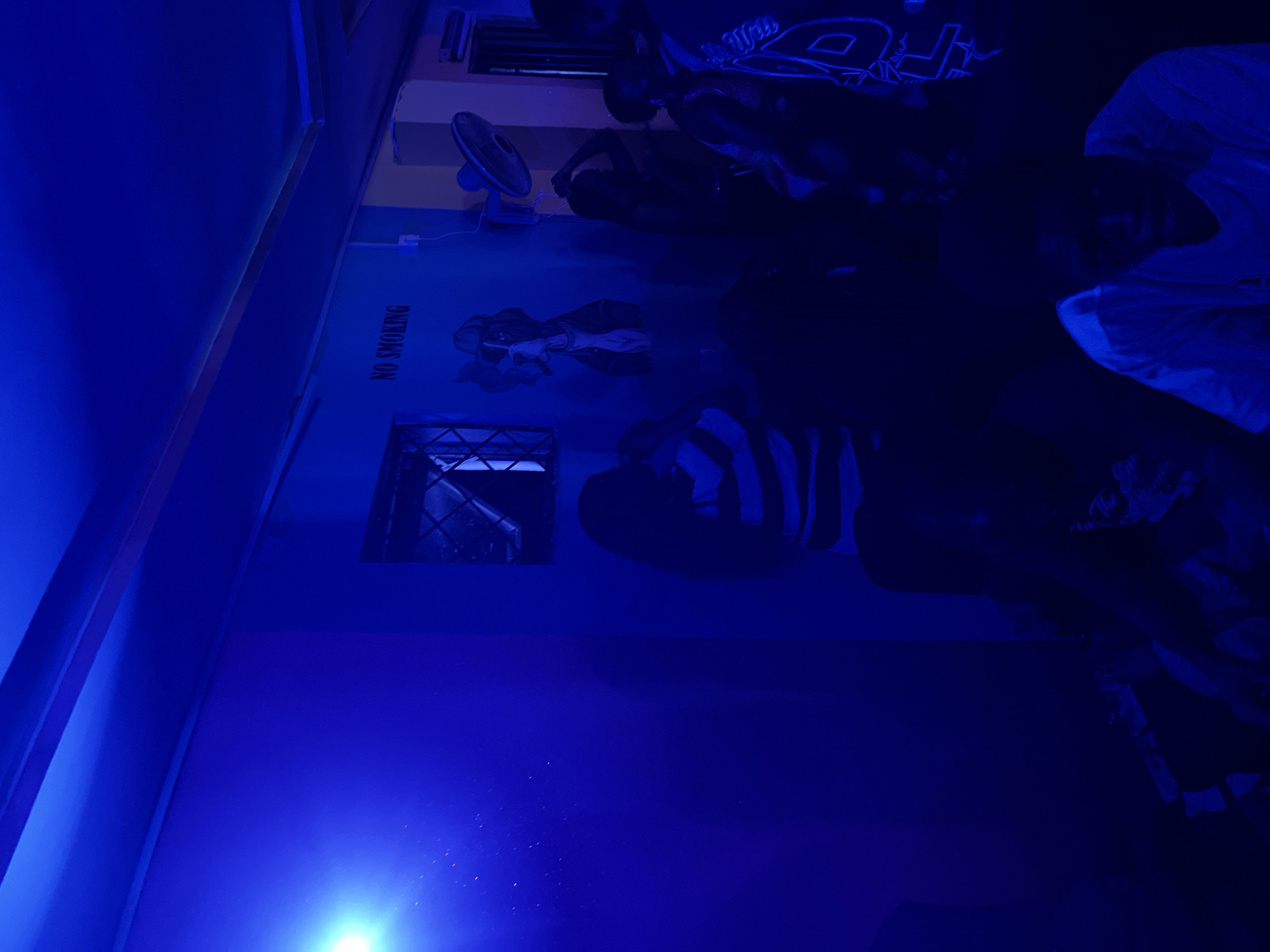 In Agwala, Nigeria, this dimly lit indoor scene is illuminated by intense blue lighting, creating a club or bar atmosphere. Several individuals are present, their features obscured by the low light and blue hue. In the foreground, a person wearing a white t-shirt is seated, looking downwards. To the left, another individual with long dark hair, wearing a striped shirt, is also seated. In the background, more people are visible, some standing and interacting. A mural depicting a figure in a hat and a "NO SMOKING" sign are visible on the blue-painted wall. A fan is mounted on the wall near the mural. The environment appears somewhat hazy, possibly due to smoke or lighting effects.