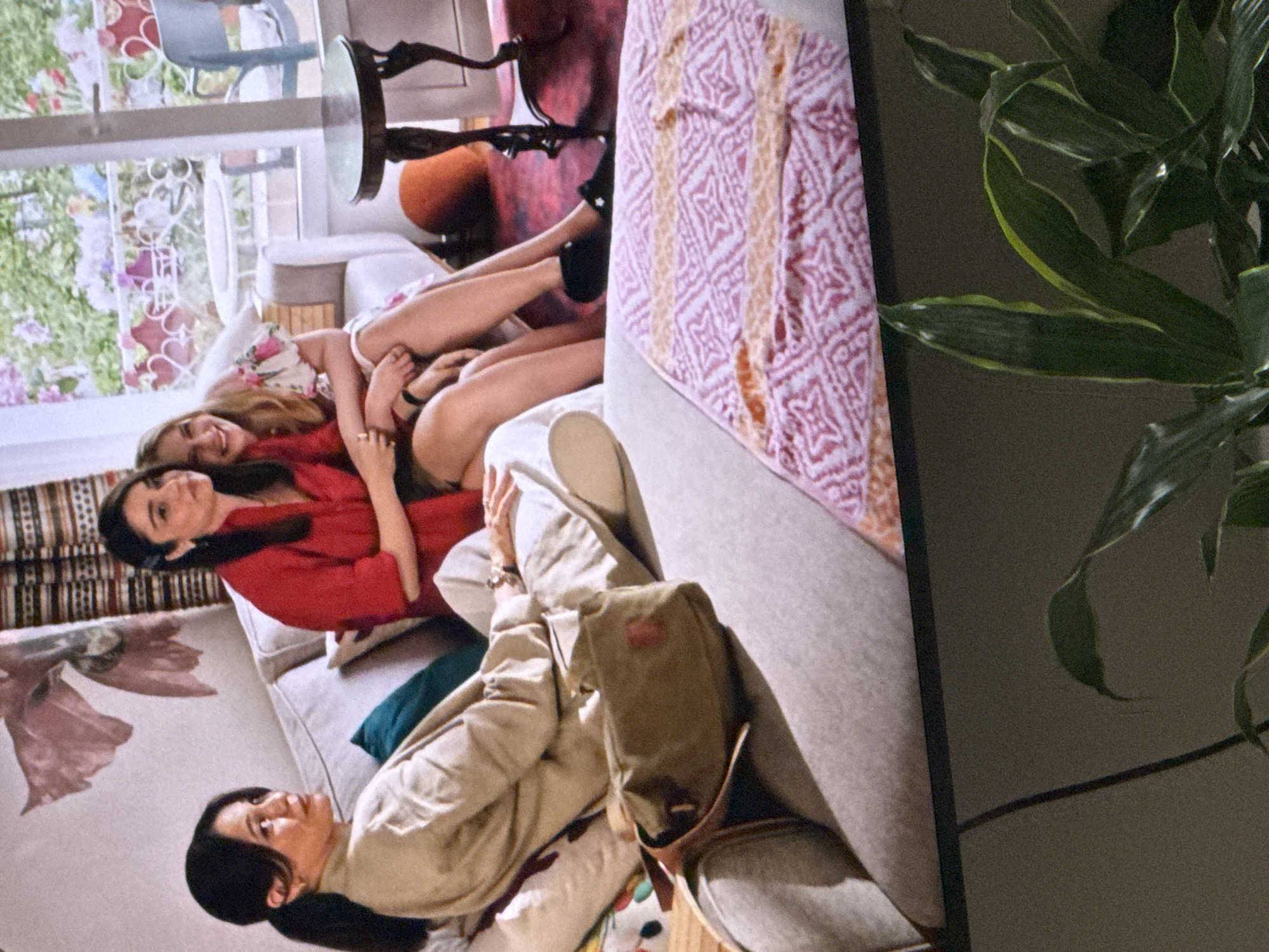 Three people are seated on a sofa in what appears to be a living room. A woman with dark hair pulled back in a ponytail is seated on the left, wearing a tan jacket and beige pants, with a green crossbody bag resting on her lap. Next to her, a woman in a red top is seated, facing away from the camera. Between them, a younger woman with blonde hair, wearing a floral print dress, is seated and leaning on the woman in the red top, smiling and looking towards the camera. Behind them, a large window offers a view of an outdoor area with plants and furniture. The sofa is adorned with cushions and a patterned blanket. In the foreground, a houseplant is partially visible, obscuring the bottom of the image.