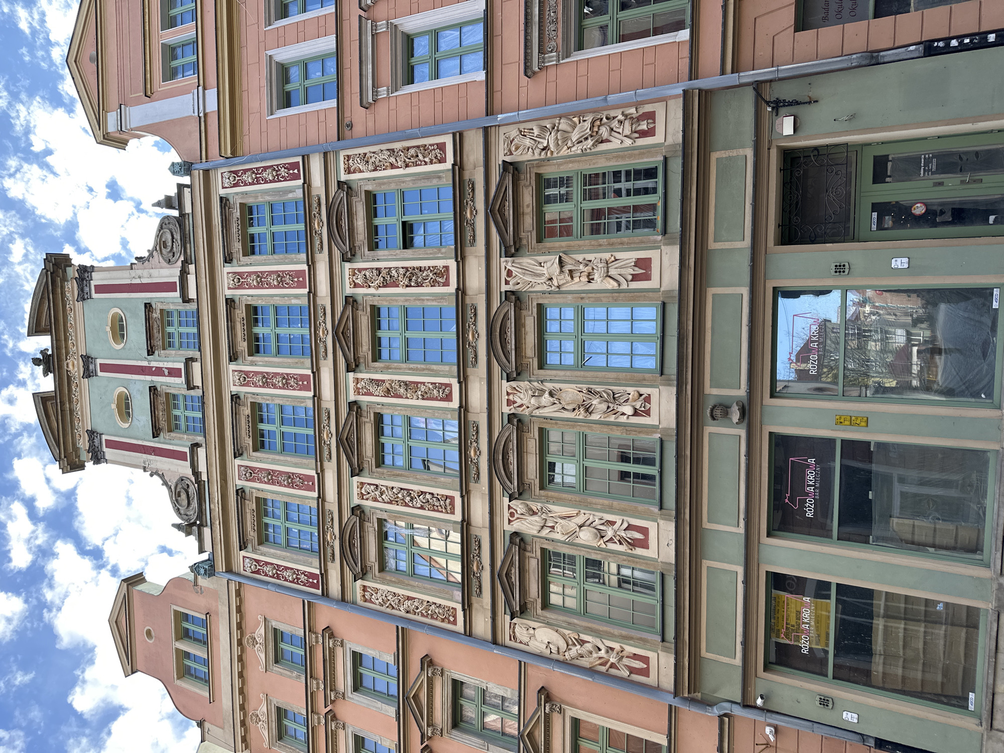 Here's a detailed description of the image:
The image shows a series of colorful, ornate buildings in what appears to be a historic district. The buildings have multiple stories, with rows of windows that reflect the blue sky. The architectural style is characterized by detailed moldings, decorative carvings, and various color schemes, including pink, beige, green, and red accents. The building on the right has ground-level storefronts with large windows, displaying signs and reflections of the street scene. One visible sign reads "RÓŻOWA KROWA BAR MLECZNY". The photograph is taken from a ground-level perspective looking upward, showing the buildings in detail, with a portion of the sky visible. The location is likely in the city of Wrocław, Poland, given the architectural style of the buildings and the sign in Polish.