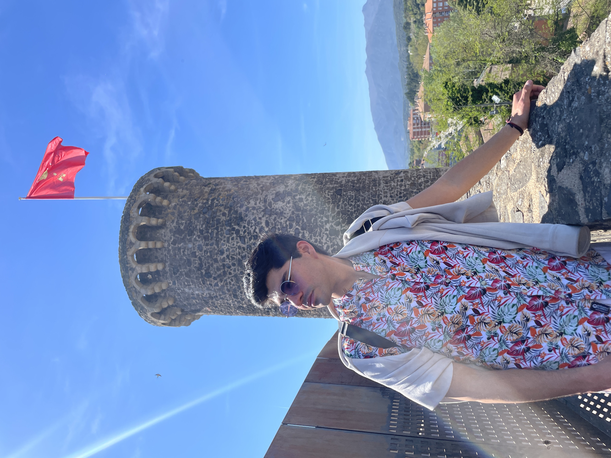 A medium shot features a man in a floral shirt and sunglasses, leaning against a stone structure with his arm extended towards a stone wall. He has a light-colored jacket draped over his shoulder and a dark strap across his body. The stone structure, identified as a tower of the medieval castle in Hostalric, Spain, is to the left and partially behind him. A red flag with a coat of arms flies atop the tower. The background includes a view of rolling hills and buildings. The sky is blue, and a bird is visible in the upper left. There are bright light flares in the lower left corner of the image.