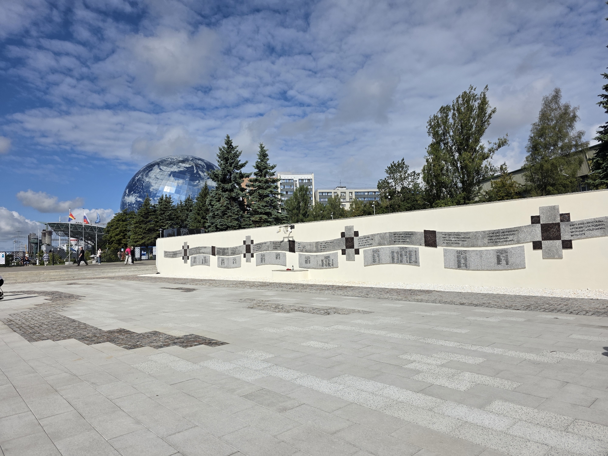 The image depicts a wide, paved plaza in front of a curved wall adorned with numerous plaques. Behind the wall, evergreen trees are visible, and further in the background, modern buildings and a large geodesic dome structure, which resembles the Earth, stand against a partly cloudy sky. The plaza is paved with large, light-colored square tiles with some areas featuring smaller, darker square inlays arranged in a zig-zag pattern. People are scattered across the plaza and walking on steps leading up to a building to the left, which features flags. The wall itself is cream-colored and features a series of dark, square elements interspersed with curved, light-colored plaques bearing text, presumably names or historical records. Two small red objects, possibly candles, are placed on a shelf beneath some of the plaques. The scene is likely a public memorial or monument, given the nature of the wall and plaques. This location is the "Monument to the Heroes of the Great Patriotic War" in Kaliningrad, Russia, situated near the "World Ocean Museum" which houses the geodesic dome.