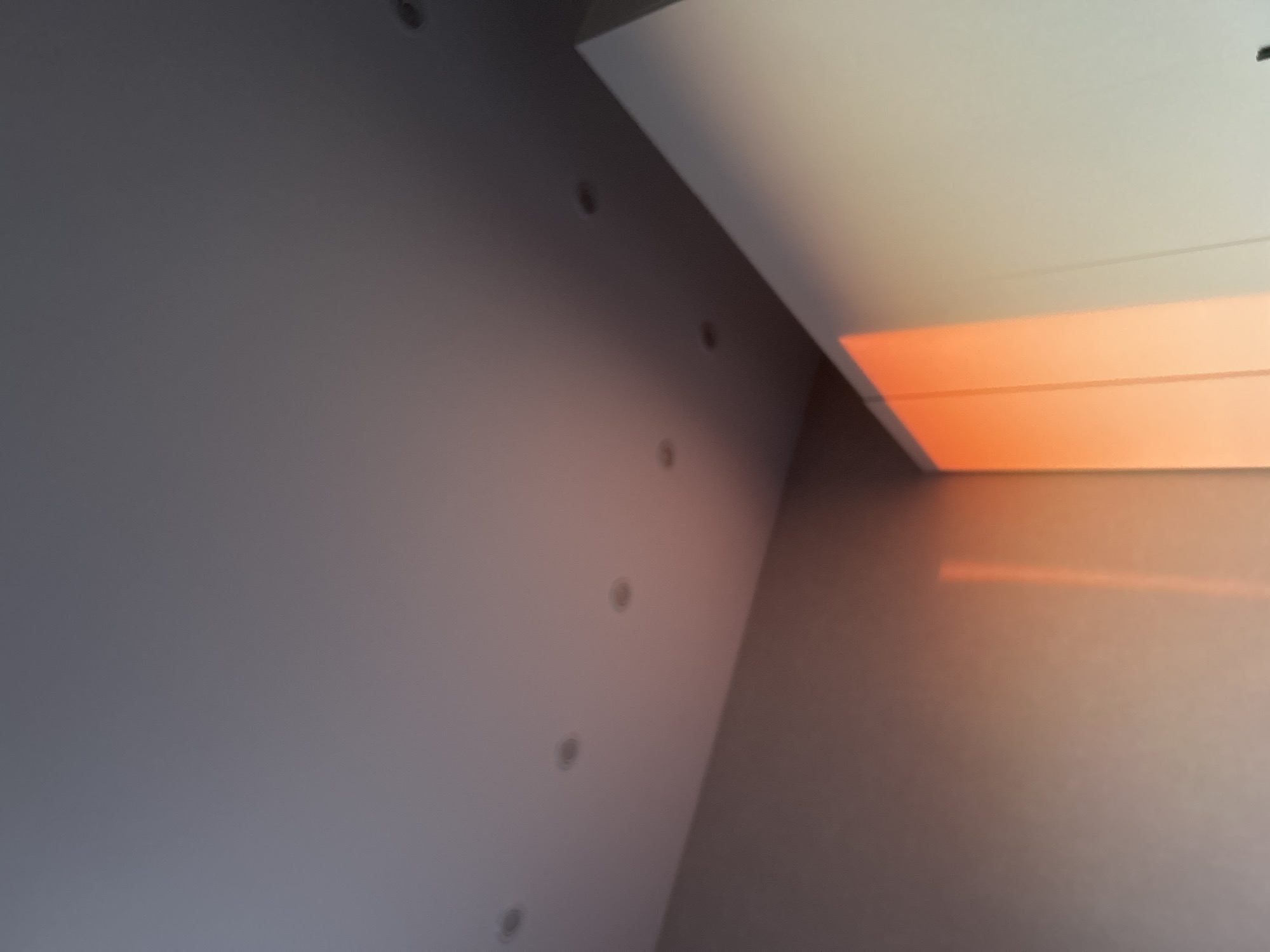 Here is a description of the image:
The image shows an interior scene, likely a room in a building. The ceiling is white, and there is an orange-glowing light fixture or opening in the upper right area of the image. The walls are gray and feature a vertical row of small, circular indentations or possibly light fixtures. The photo appears to be taken at an upward angle, capturing the intersection of the wall and ceiling. There is no readily identifiable location information from the image. No people, objects, or actions are discernible beyond the described features of the room.