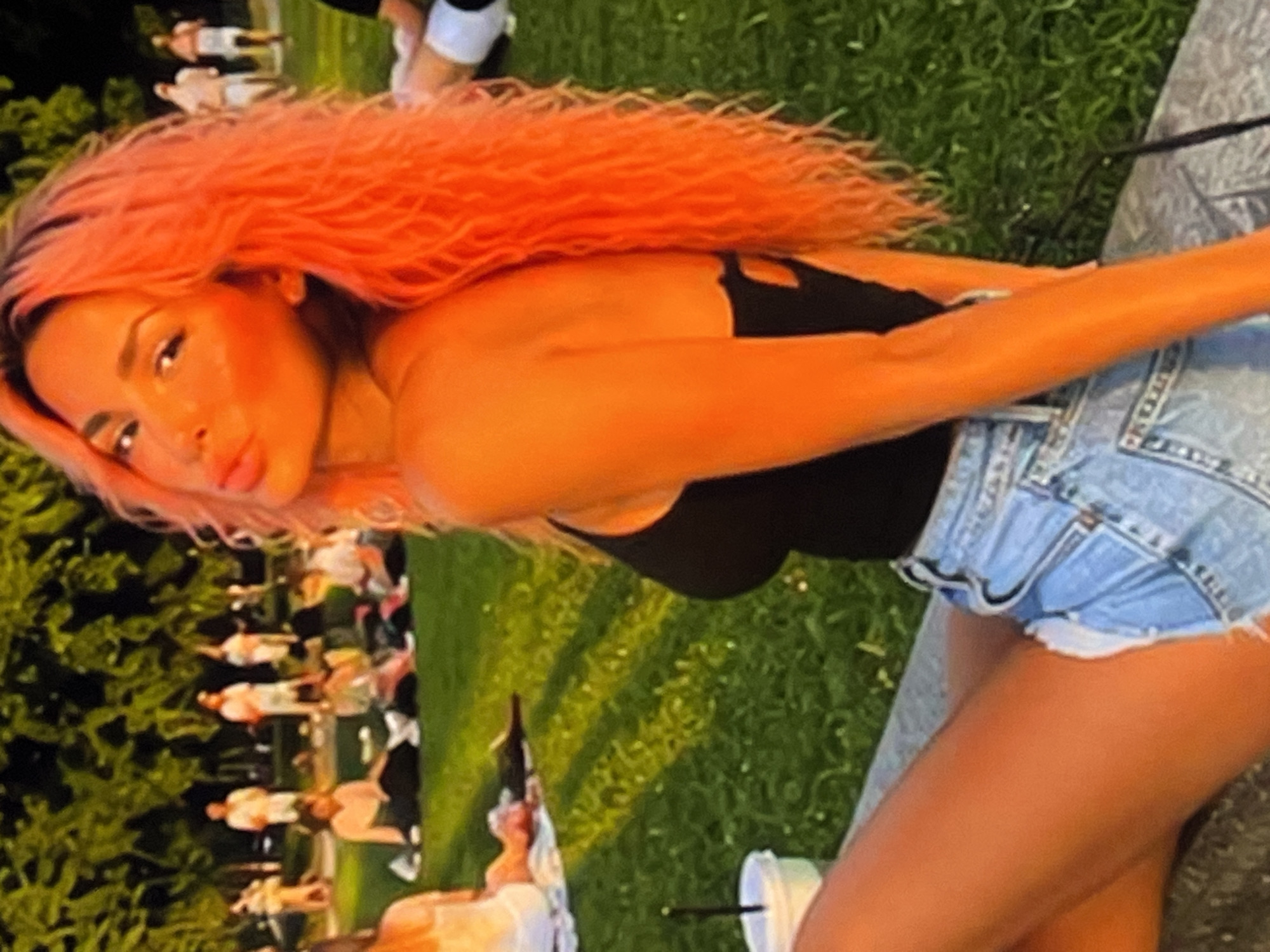 In an outdoor park setting under dappled sunlight filtering through trees, a young woman with bright orange, curly hair is the primary subject. She is seated in the foreground, leaning back slightly with her head turned towards the viewer, her lips slightly parted. She is wearing a black tank top and light-wash denim shorts. Her arms and legs are visible, and her skin is tanned.
Behind her, other individuals are spread out on a grassy area, some seated on blankets, others standing. The people in the background are less distinct due to the focus on the foreground subject and the lighting conditions. The environment suggests a relaxed, recreational atmosphere, likely during daytime. The overall color palette is warm, dominated by the vibrant orange of the woman's hair and the green of the grass, with touches of blue from the denim.