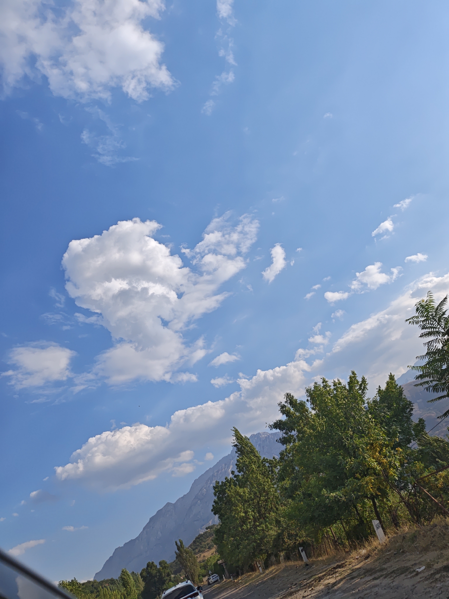 The image shows a scenic view of the sky and mountains, with trees and a road in the foreground. The sky is a clear blue, with scattered white clouds. The mountains are rugged and majestic, with a hint of snow on their peaks. The trees are lush and green, with some of them showing signs of autumn colors. The road is paved and appears to be well-maintained. The scene is peaceful and serene, evoking a sense of tranquility and natural beauty. The location is in Brichmulla, Uzbekistan, a popular tourist destination known for its stunning natural landscapes.