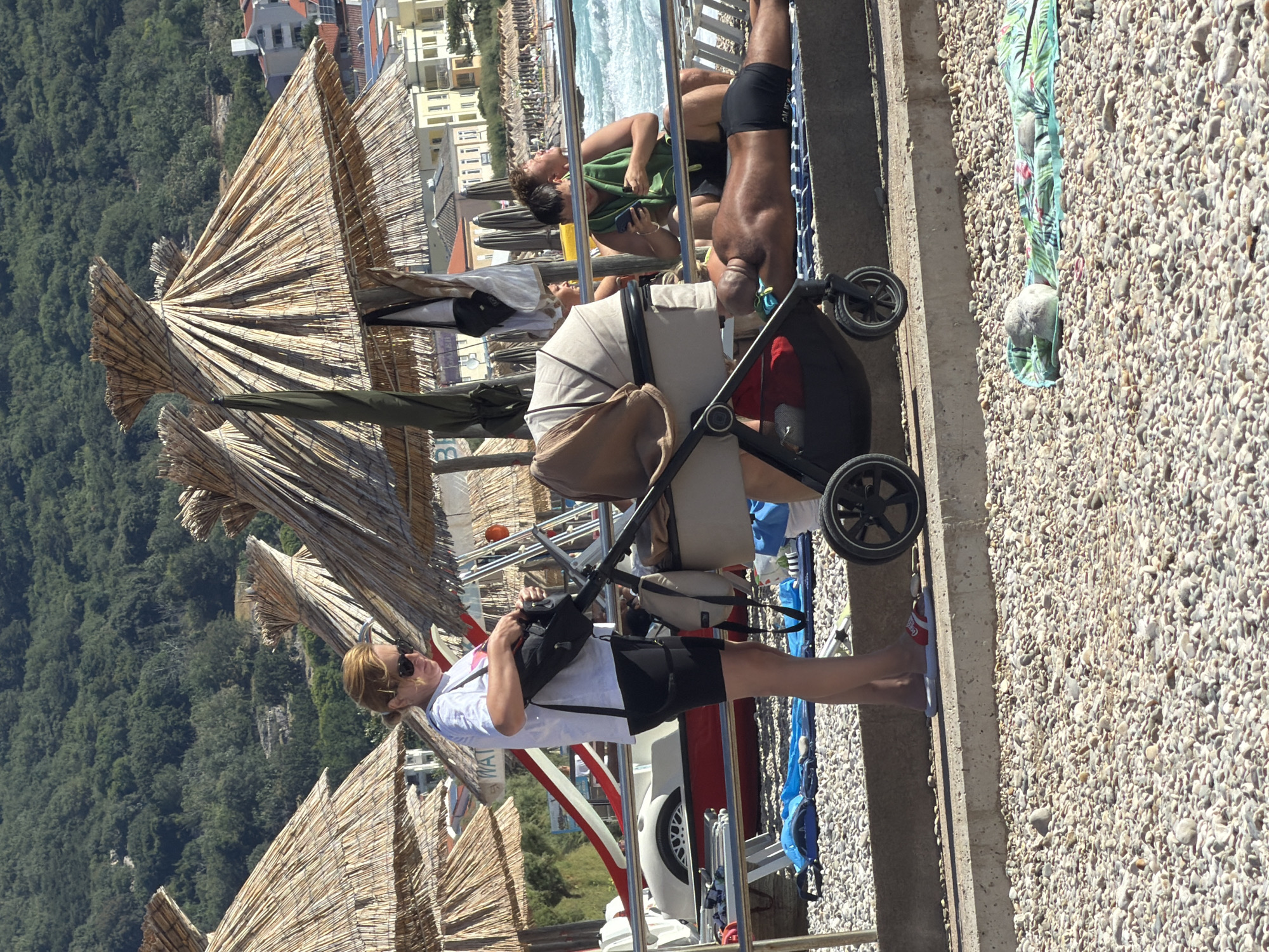 In Općina Baška, Croatia, on a sunny day at a pebble beach, a woman with blonde hair pulled back, wearing sunglasses, a white t-shirt, and black shorts, is attending to a stroller. She is holding a black bag. The stroller is a beige bassinet type with black wheels. Behind the woman, a man in a green towel is sitting and looking at his phone, and another person is lying on a beach mat. The background shows the sea with waves, a shoreline lined with buildings, and a densely wooded hill. Several thatched umbrellas are set up on the beach. In the foreground, a beach mat with a tropical pattern is laid out on the pebbles.