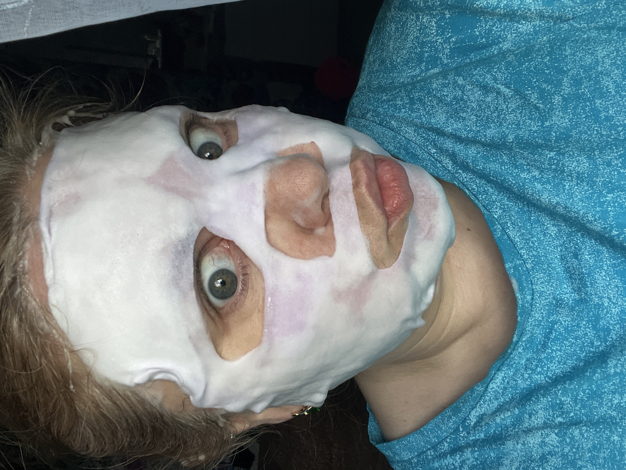 A woman wearing a white sheet mask on her face is photographed from a low angle. The mask has cutouts for the eyes, nose, and mouth. The woman has light-colored hair and greenish-gray eyes. She is wearing a blue shirt. The background is dark and partially obscured. The location of this photograph in the city of Vesyoloye, Russia, cannot be determined from the image.
